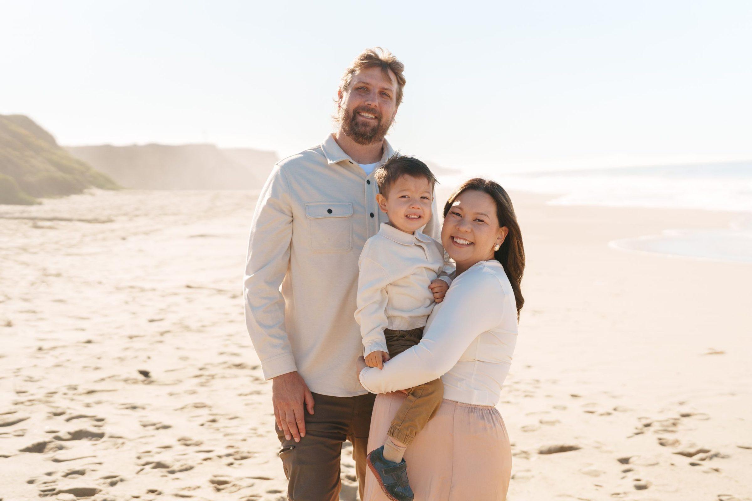 Bay Area family portrait of parents in cream tops holding their smiling toddler son on a sandy beach with coastal bluffs.