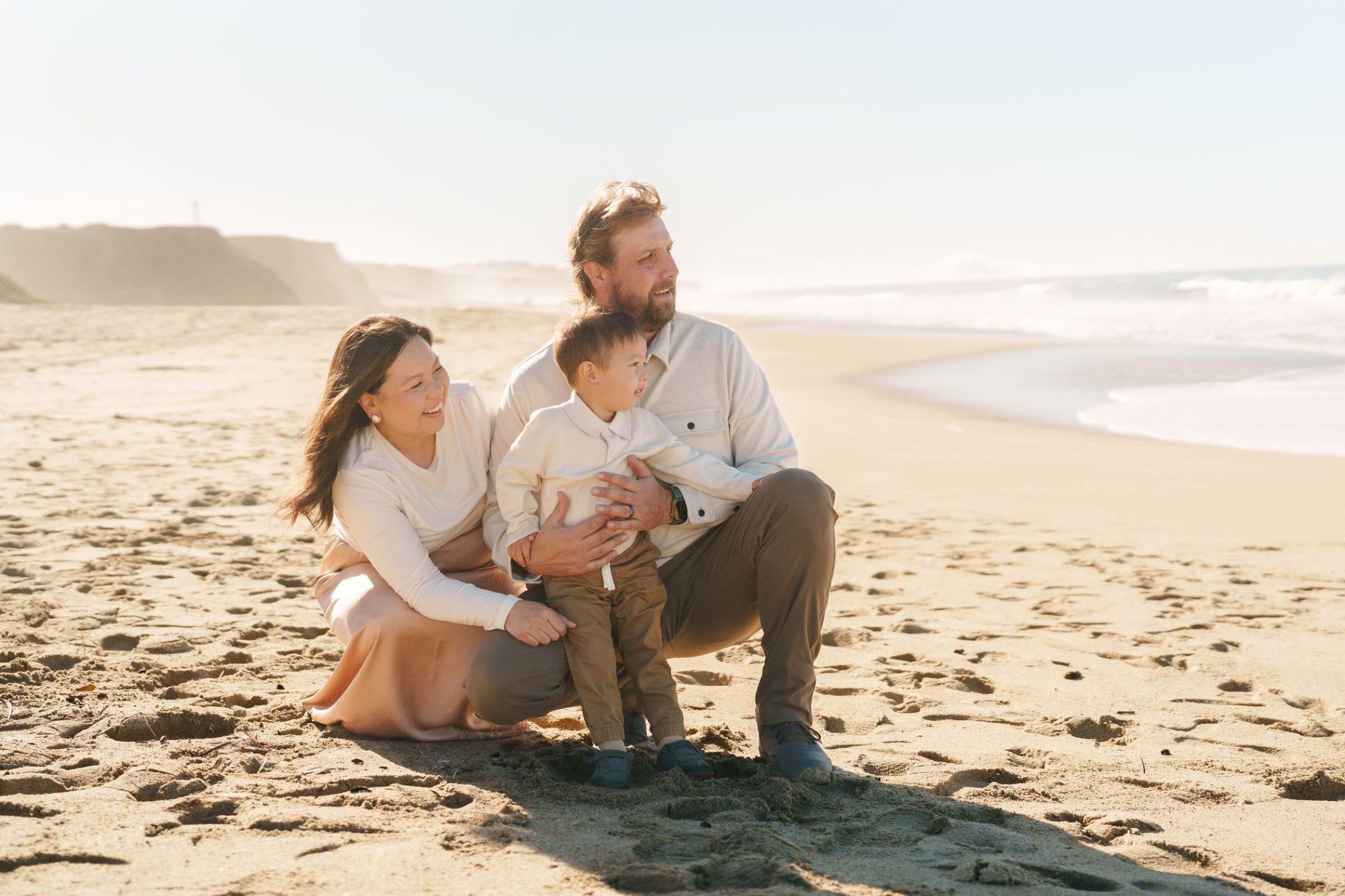 Bay Area family portrait of mom, dad and young son in cream tops on sandy beach with coastal bluffs and surf behind them