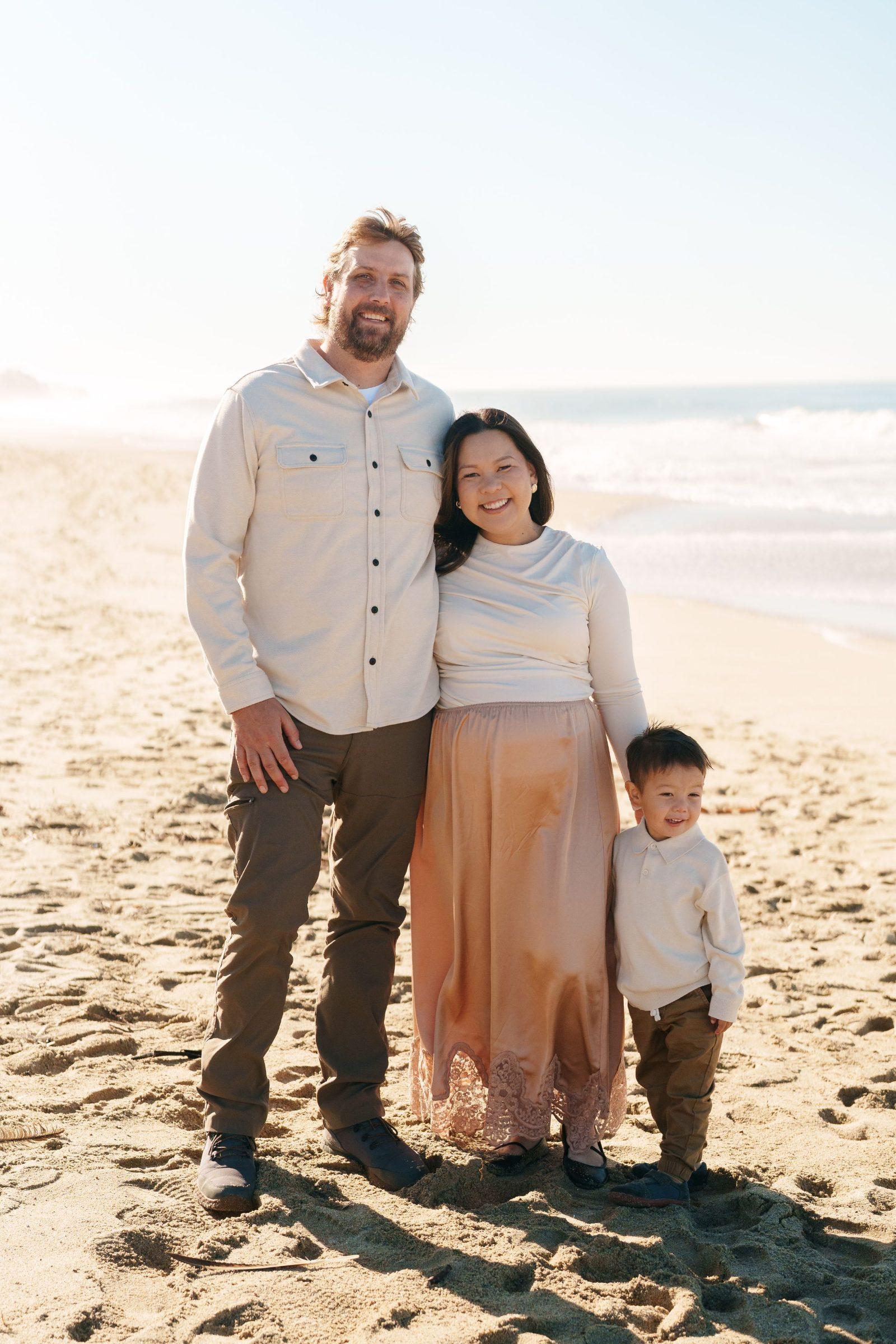 Bay Area family portrait of parents and toddler son in cream tops and peach skirt standing on sunlit sandy beach with ocean