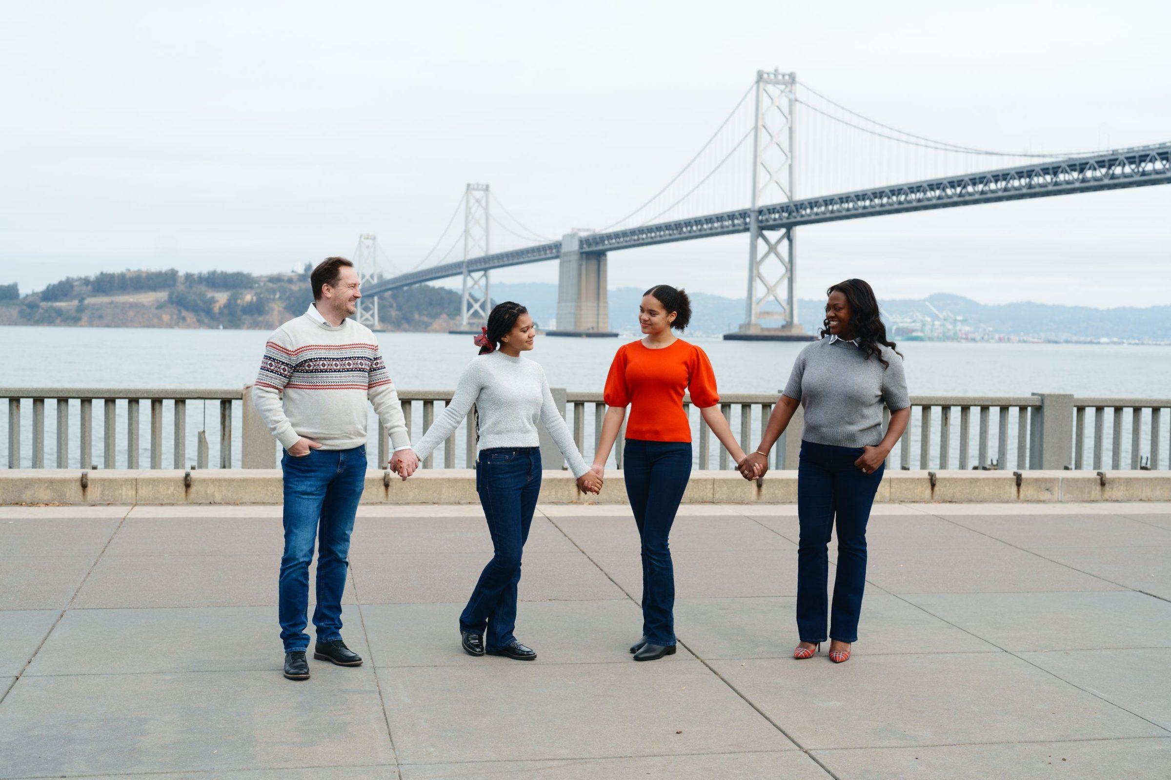 Bay Area family portrait of parents and two teen daughters holding hands along the Embarcadero with the Bay Bridge behind