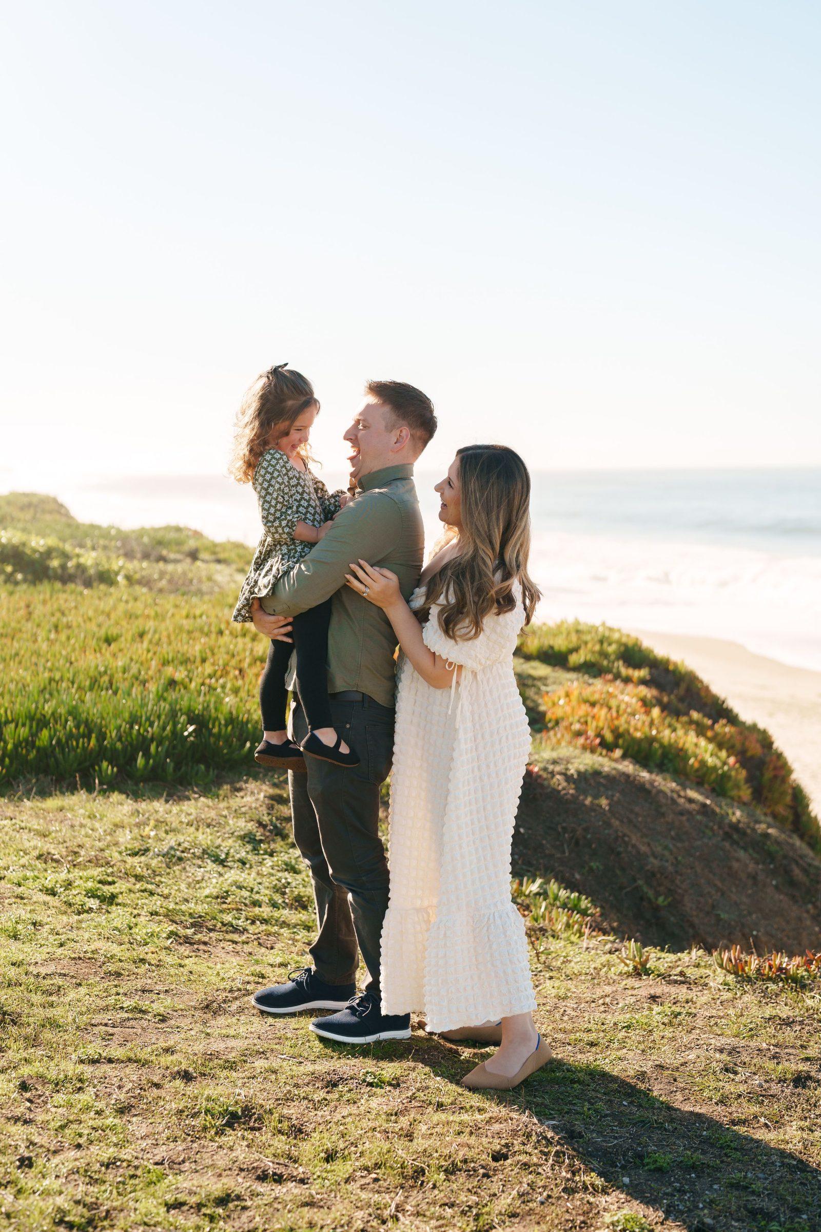 Bay Area family portrait of dad holding daughter in floral top while mom in white dress laughs beside them on a coastal