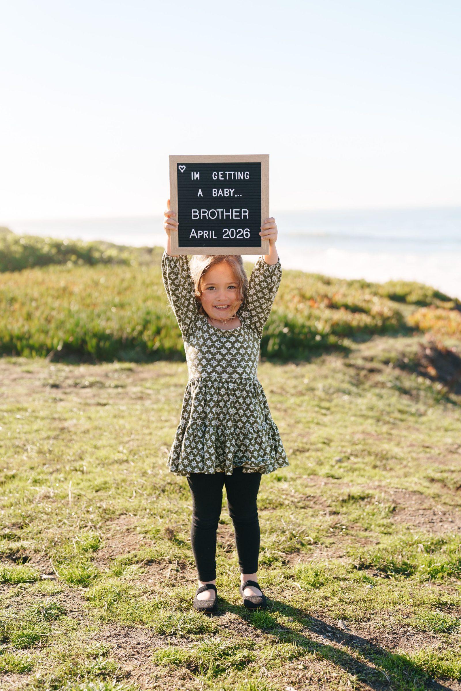 Bay Area family portrait of a young girl in a green floral dress holding a letter board announcing a baby brother coming