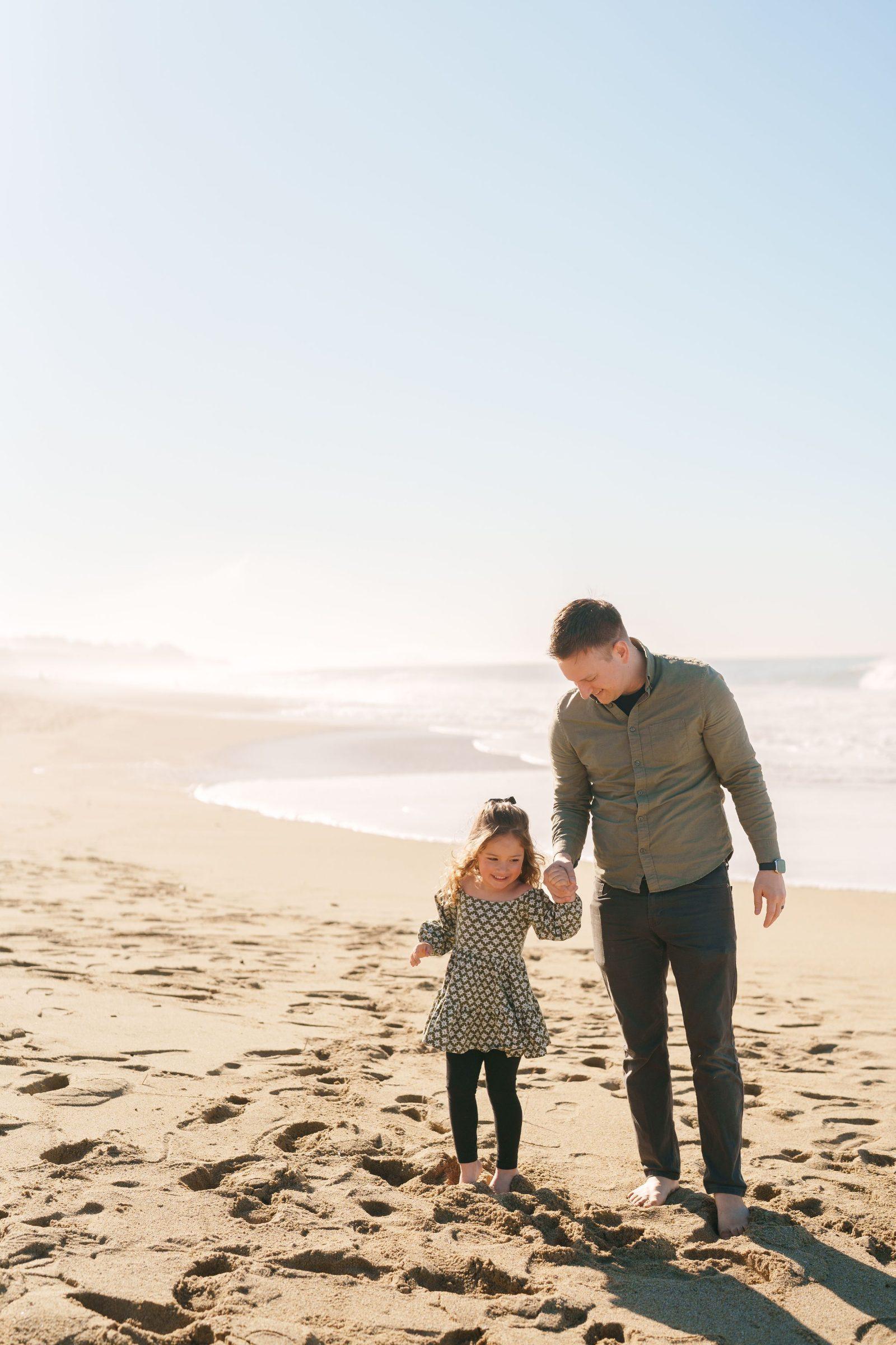 Bay Area family portrait of a dad in olive shirt holding his young daughter's hand as they walk barefoot on sunlit sand.