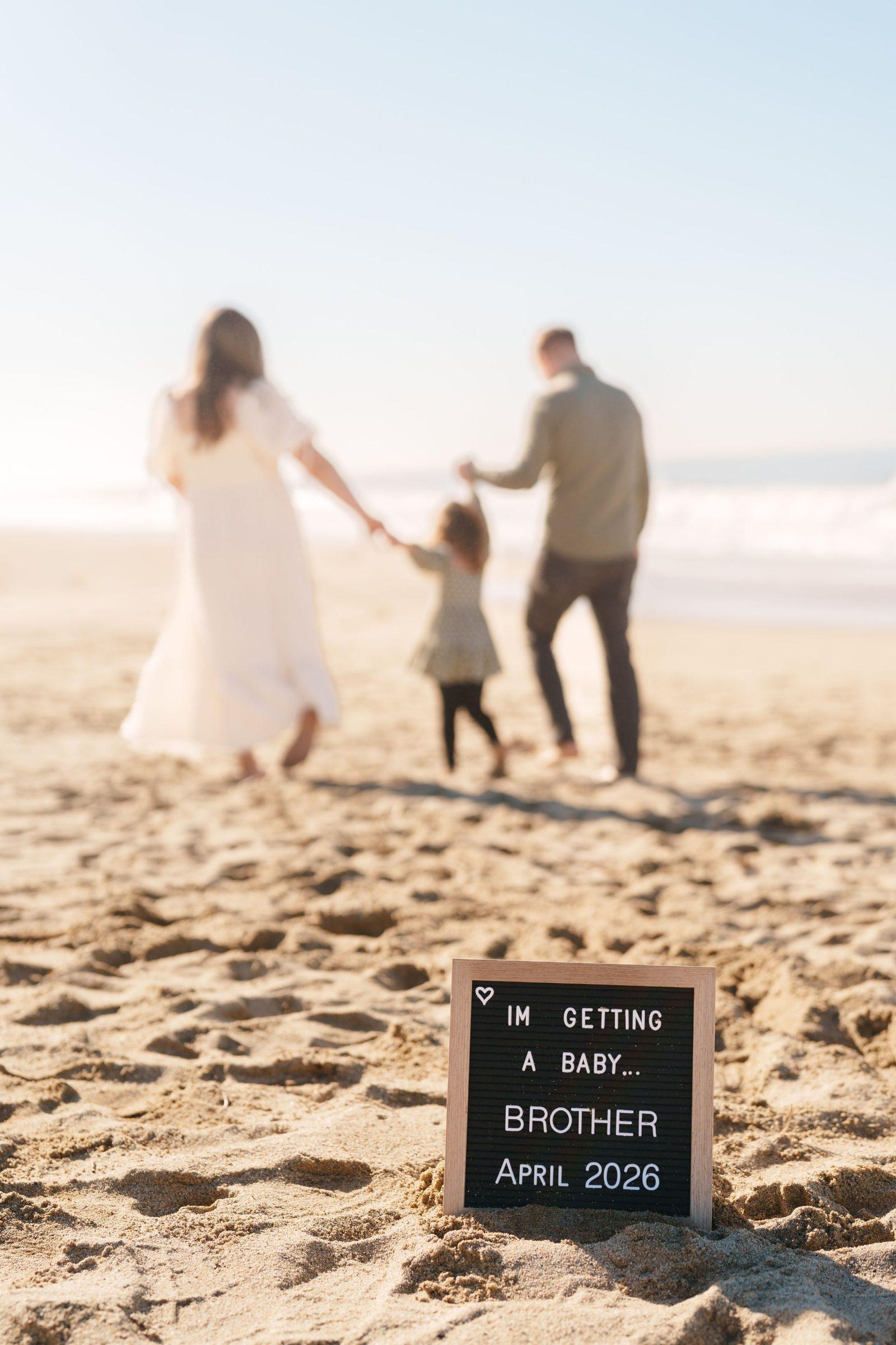 Bay Area family portrait on sandy beach with letter board announcing baby brother April 2026 as parents and toddler walk