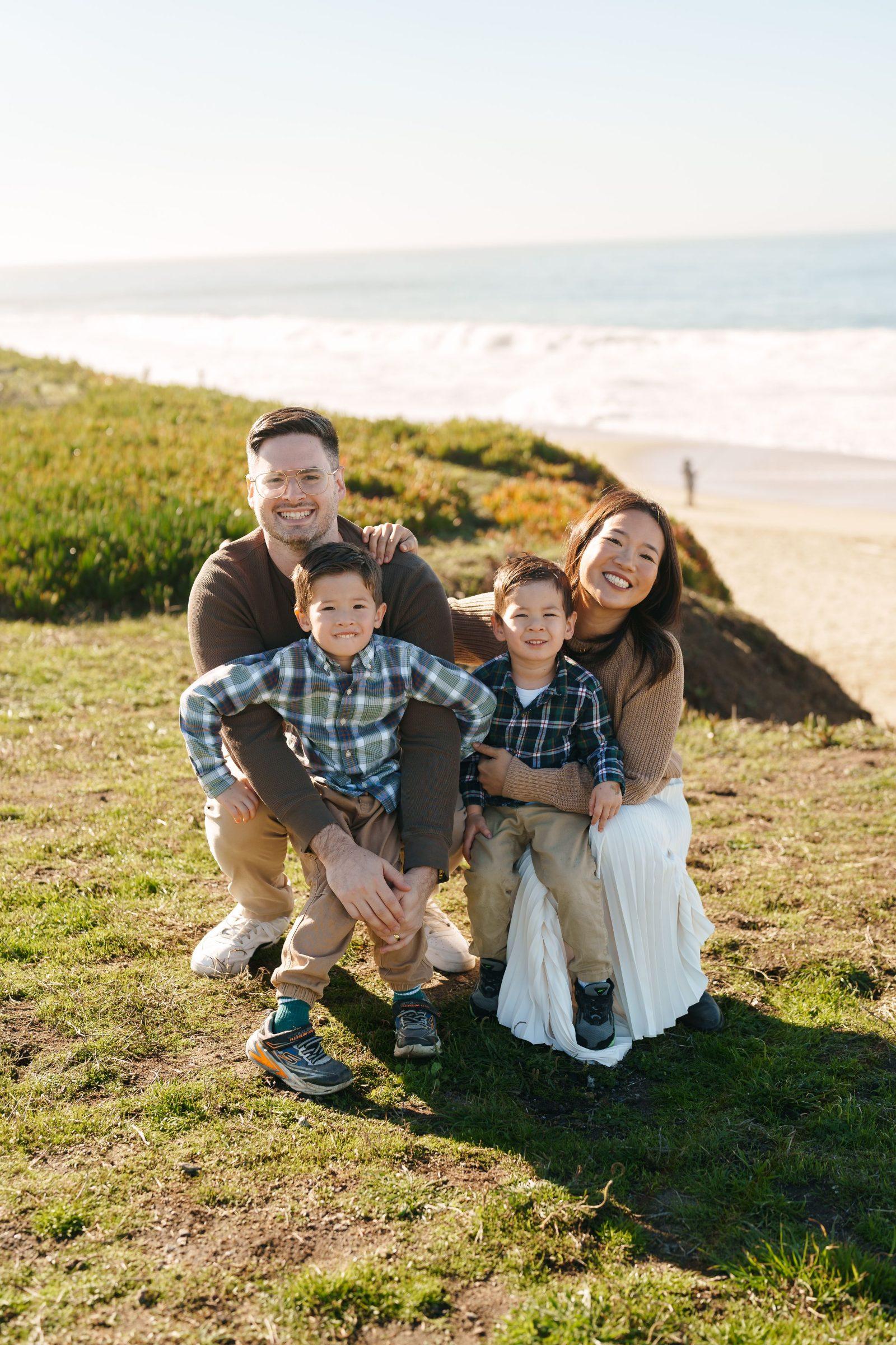 Bay Area family portrait of parents crouching on a grassy coastal bluff with two young sons in plaid shirts, ocean behind