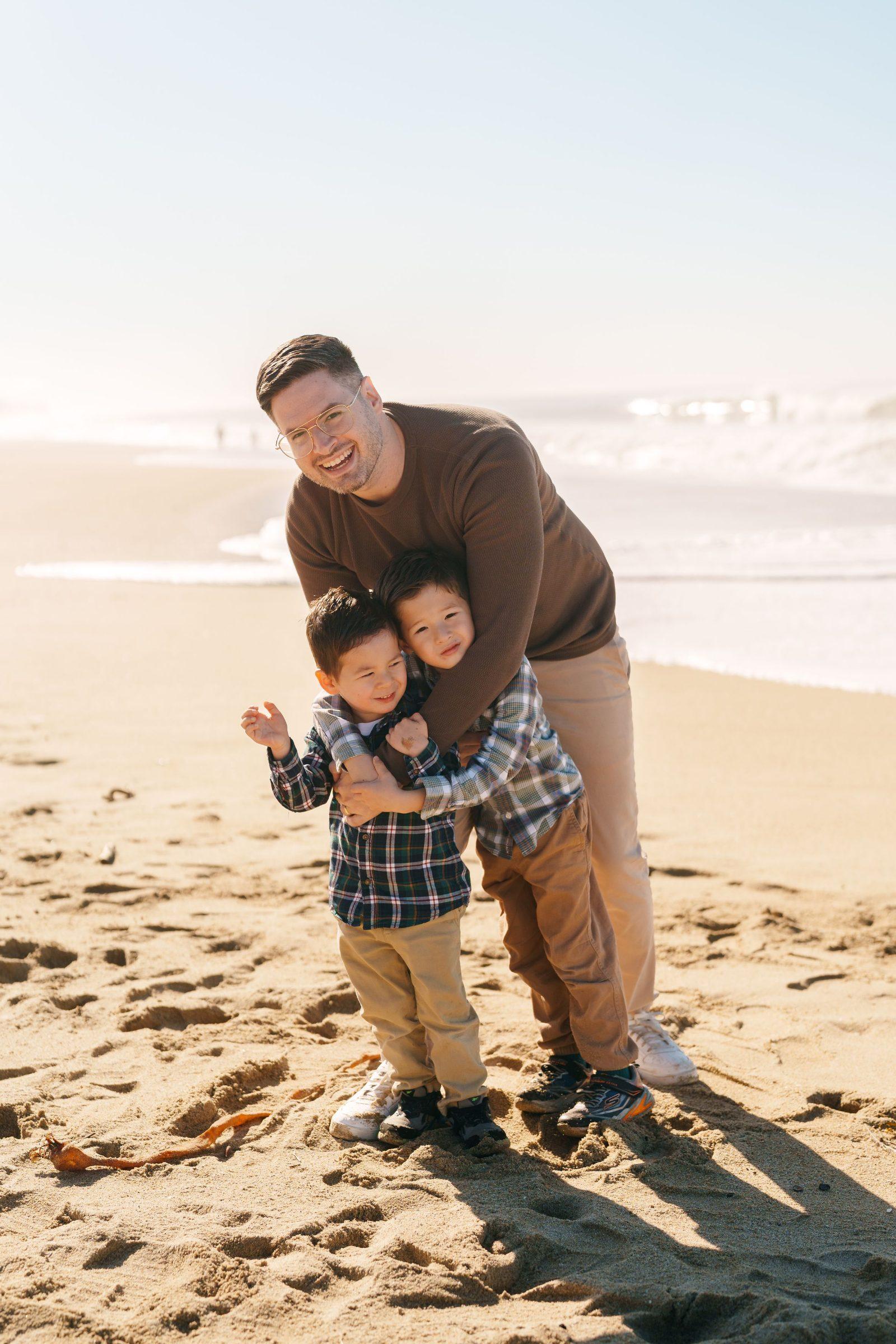 Bay Area family portrait of a dad in a brown sweater laughing while hugging his two young sons in plaid shirts on sunlit