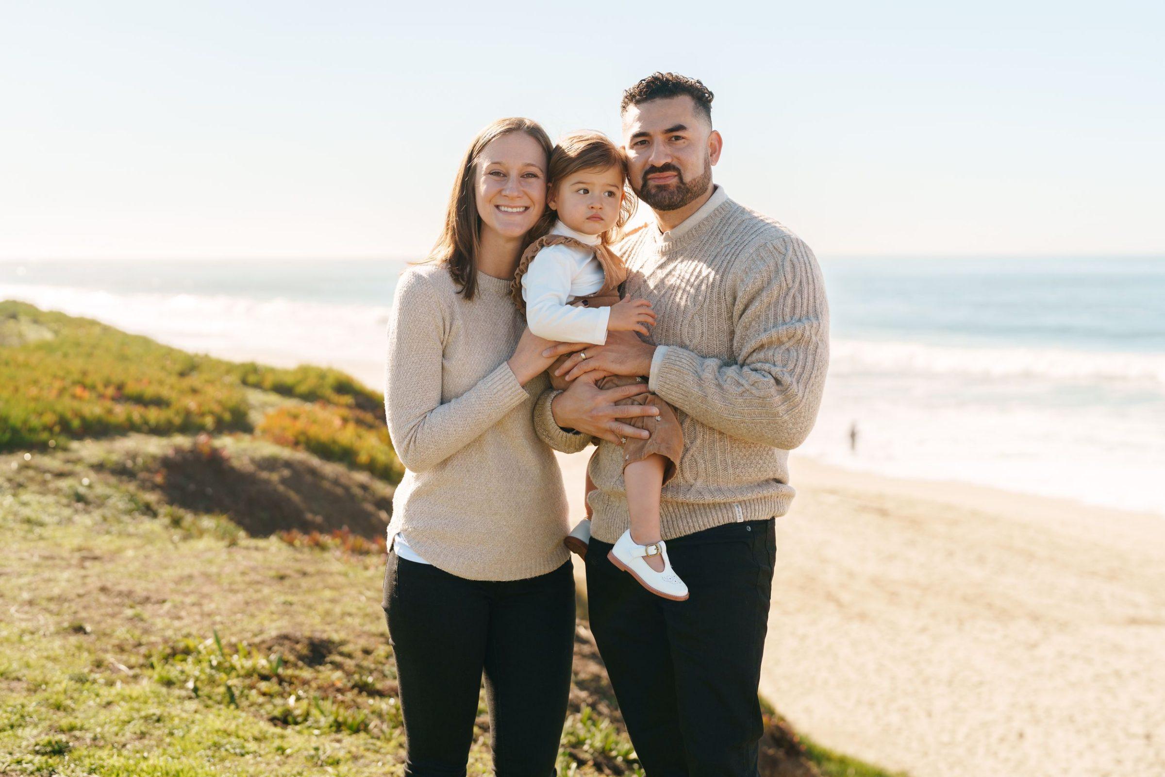 Bay Area family portrait of parents in cream sweaters holding their toddler daughter on a coastal bluff above the ocean.