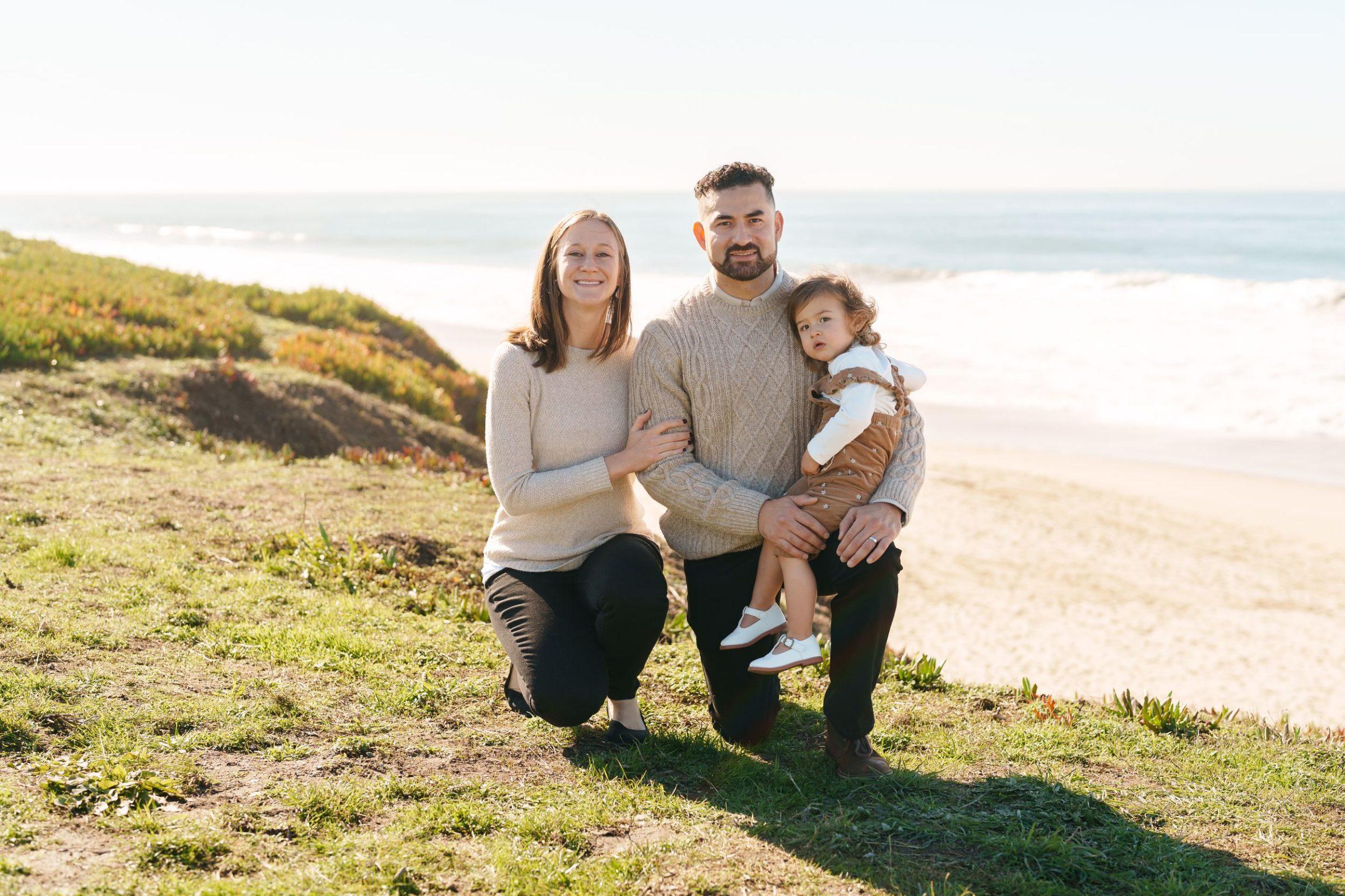 Bay Area family portrait of parents in cream sweaters kneeling on a grassy bluff, dad holding toddler daughter, ocean behind.