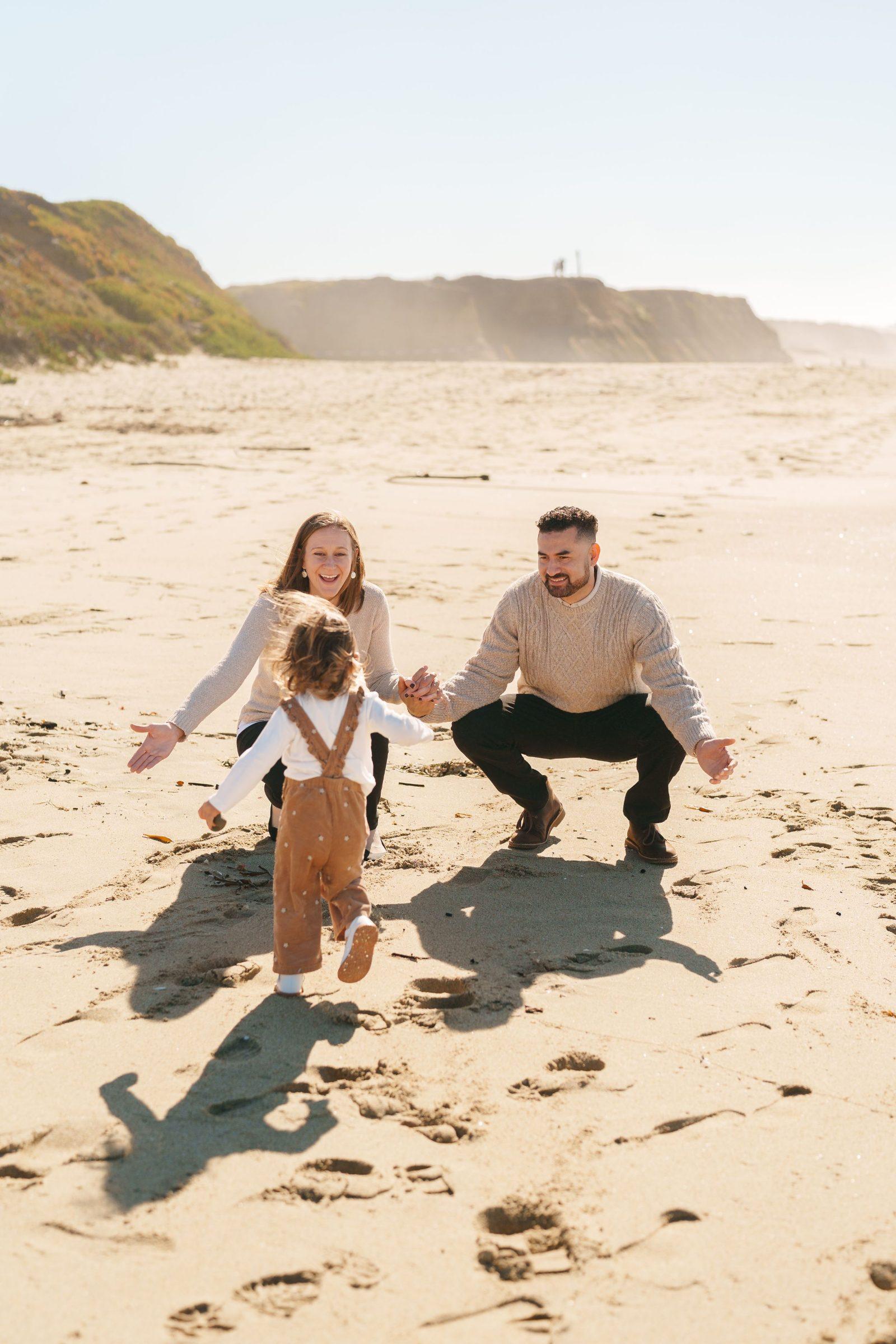 Bay Area family portrait of a toddler in brown overalls running toward her crouching parents in cream sweaters on sandy beach