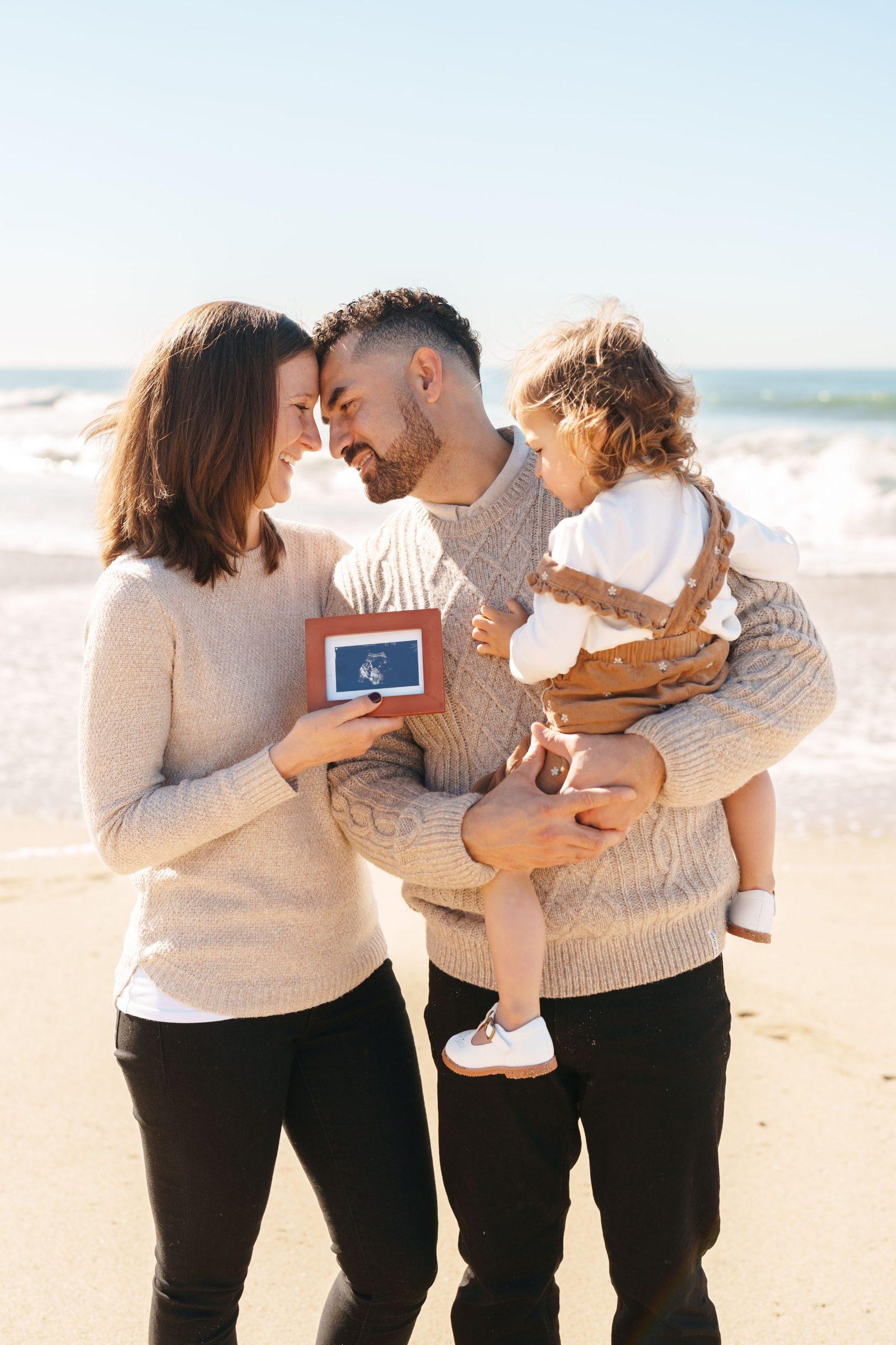 Bay Area family portrait on the beach as parents in cream sweaters touch foreheads, holding a framed ultrasound and their