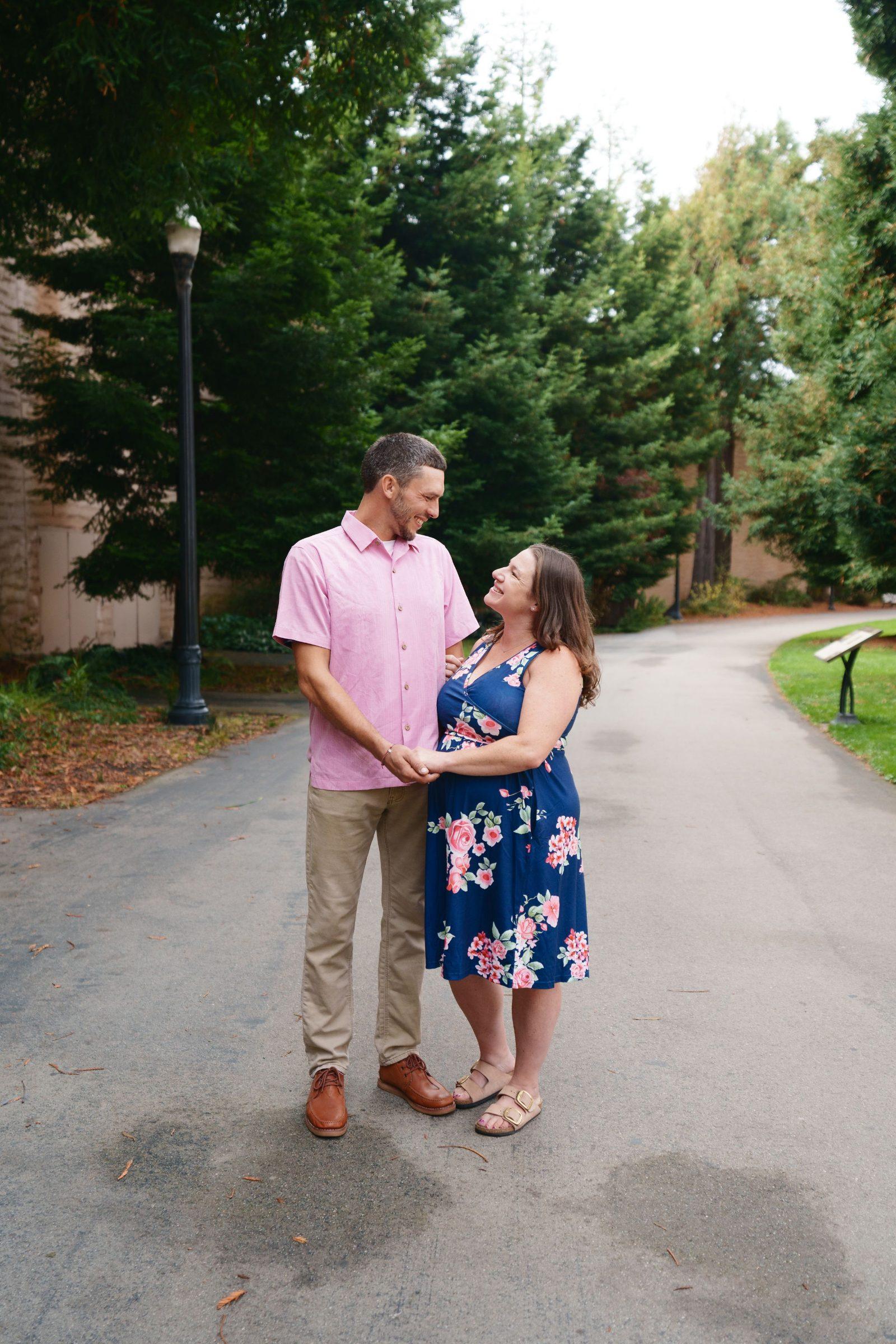 Bay Area family portrait of a couple holding hands on a park path, man in pink shirt and woman in navy floral dress smiling