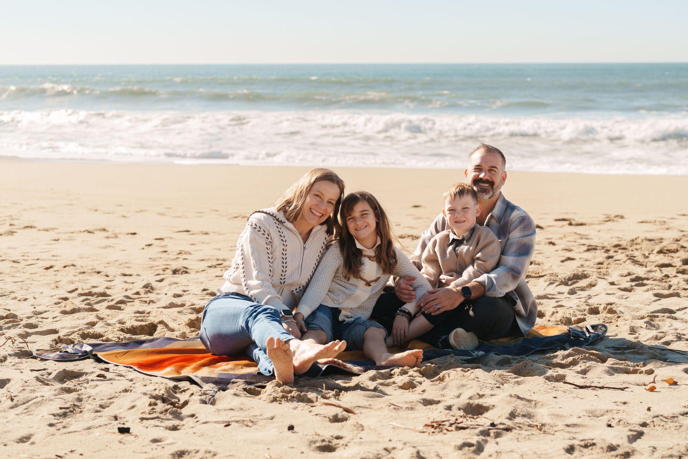 Bay Area family portrait of parents and two kids in cream sweaters sitting barefoot on a striped blanket with ocean waves