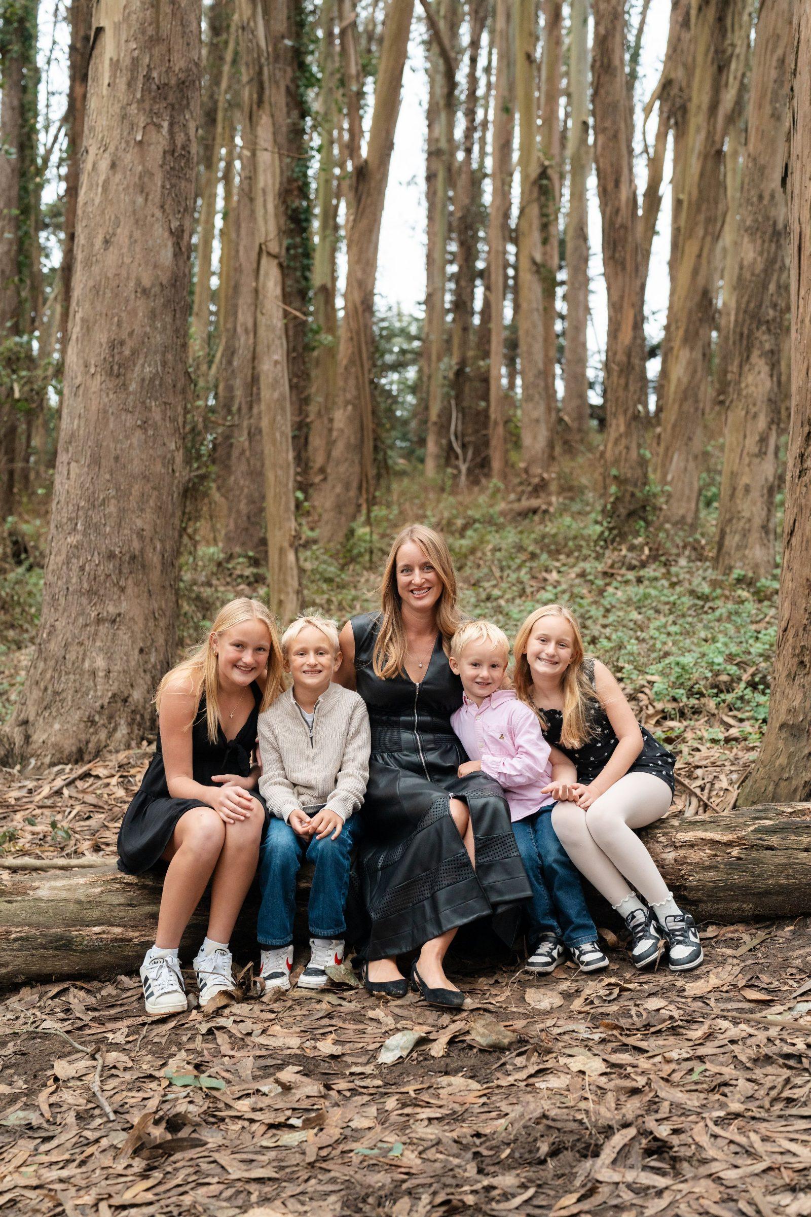 Bay Area family portrait of a blonde mother in a black dress with her four children seated on a log in a eucalyptus grove.