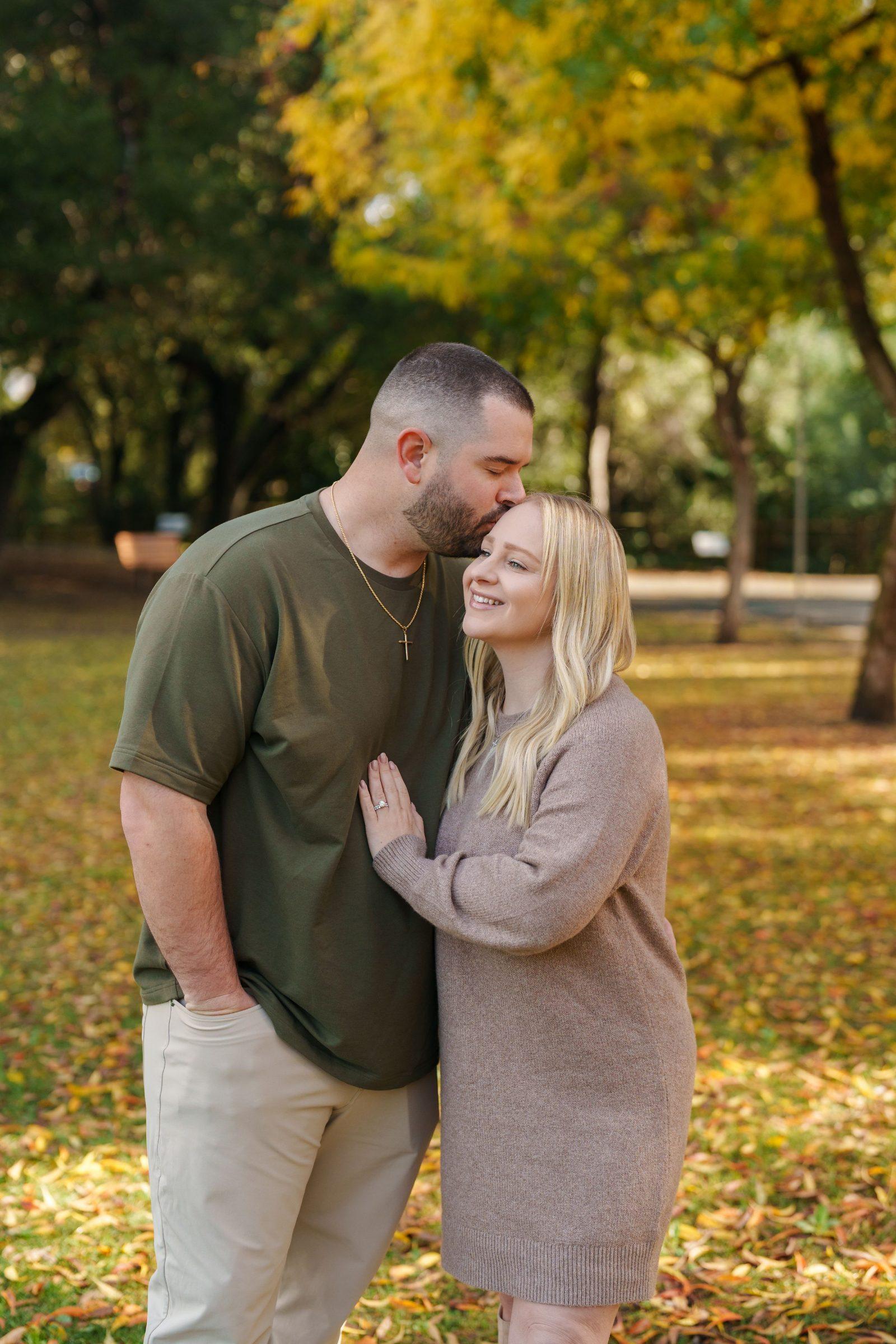 Bay Area family portrait of a husband in olive tee kissing his blonde wife's forehead in a sunlit autumn park.