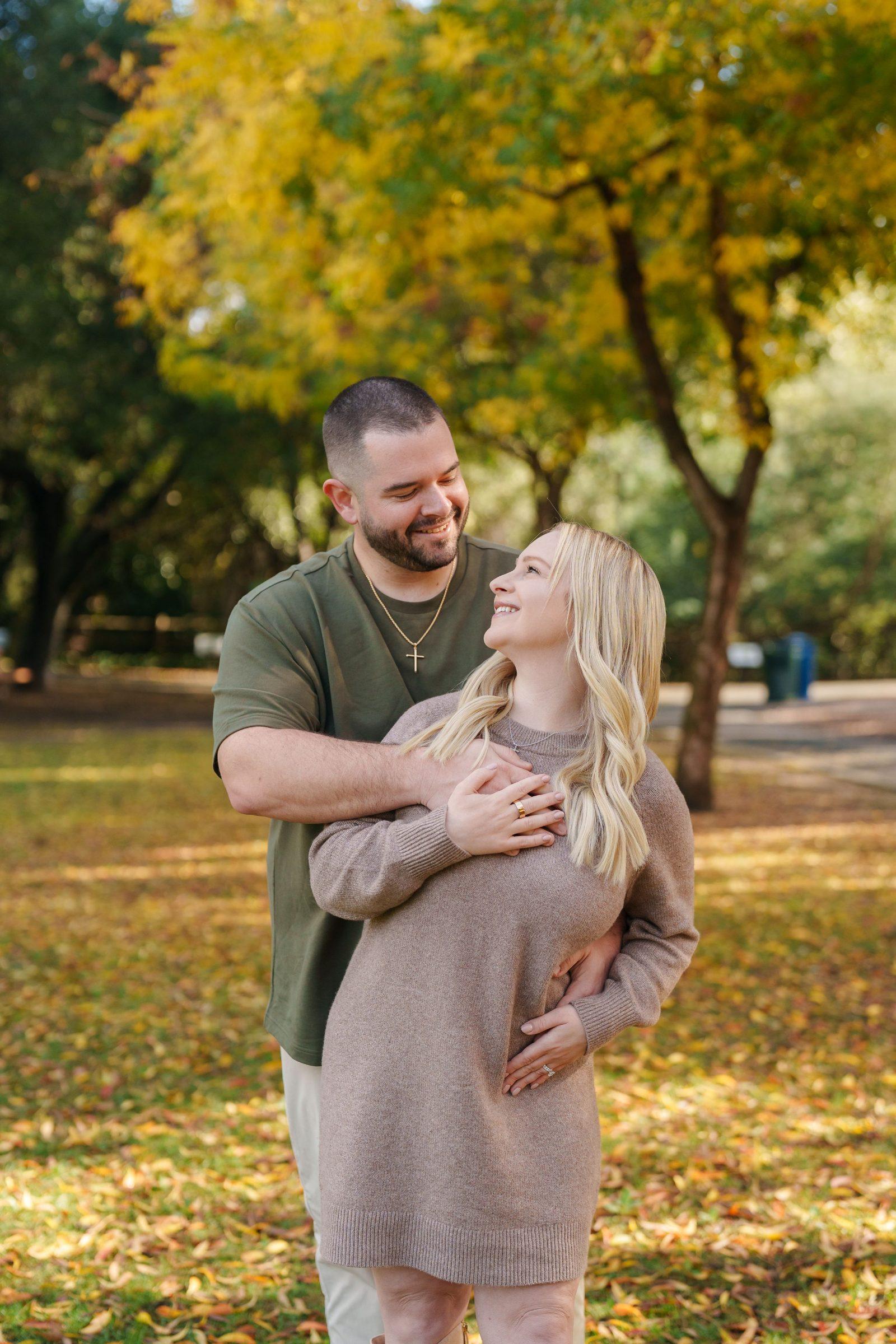 Bay Area family portrait of a couple embracing in a park, man in olive tee hugging blonde woman in taupe sweater dress among