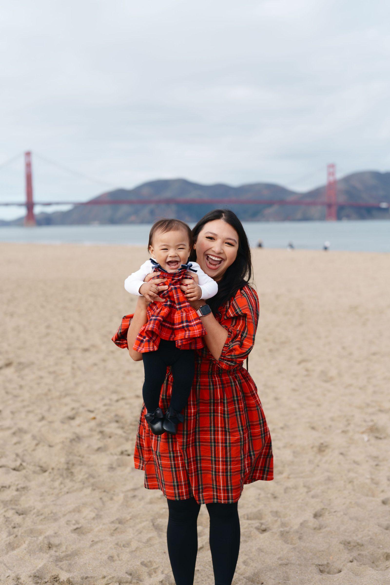Bay Area family portrait of a laughing mom holding her baby daughter, both in red tartan plaid dresses, with the Golden Gate