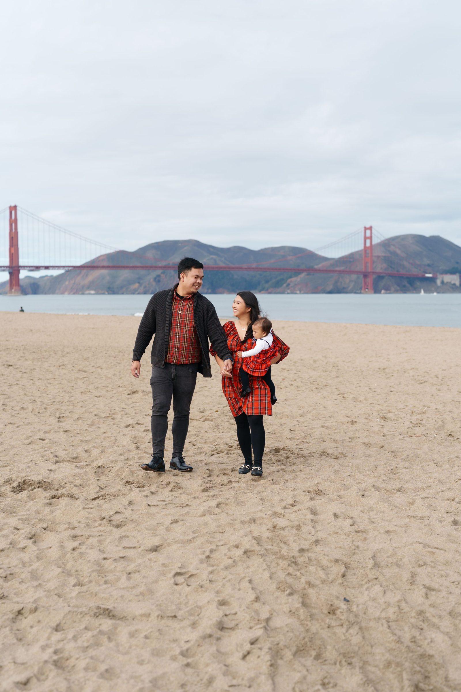 Bay Area family portrait of parents in red plaid holding their baby while walking on Crissy Field sand with the Golden Gate