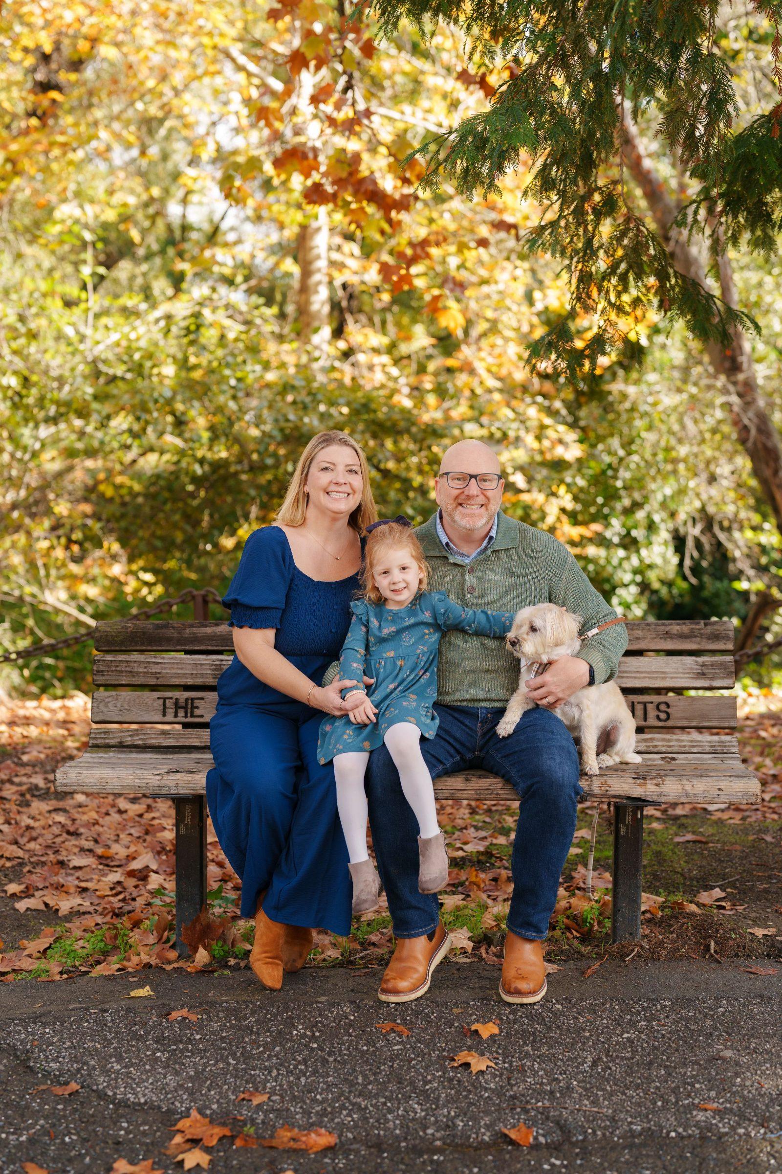 Bay Area family portrait of mom in blue dress, dad in green sweater, red-haired daughter and small dog on a park bench among