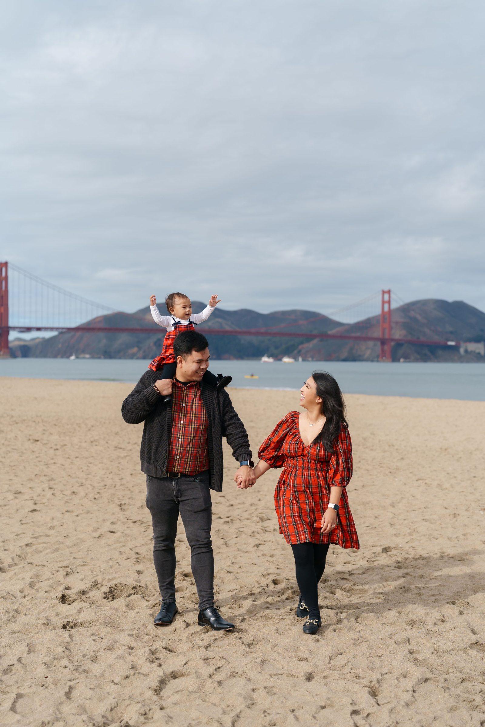Bay Area family portrait of parents in red plaid holding hands on Crissy Field sand, toddler cheering on dad's shoulders,