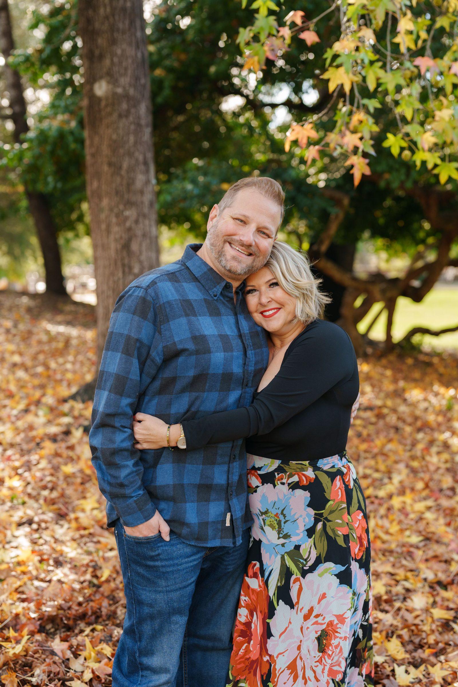 Bay Area family portrait of a couple hugging among autumn leaves, man in blue plaid shirt, woman in black top and floral