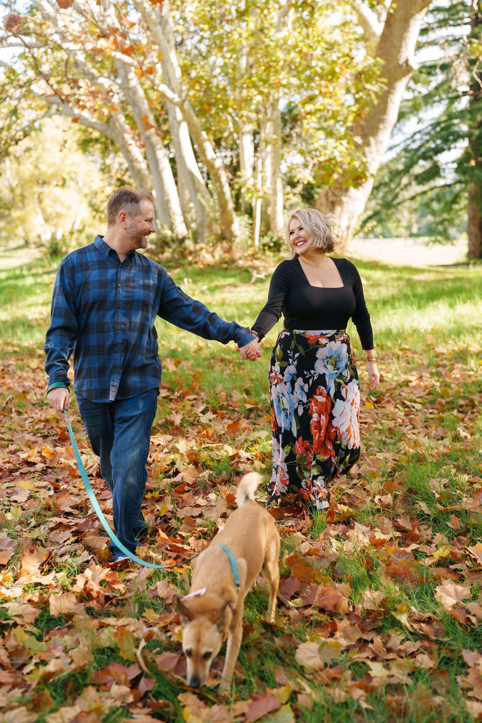 Bay Area family portrait of a couple holding hands walking their tan dog on a leash through autumn leaves under sycamore