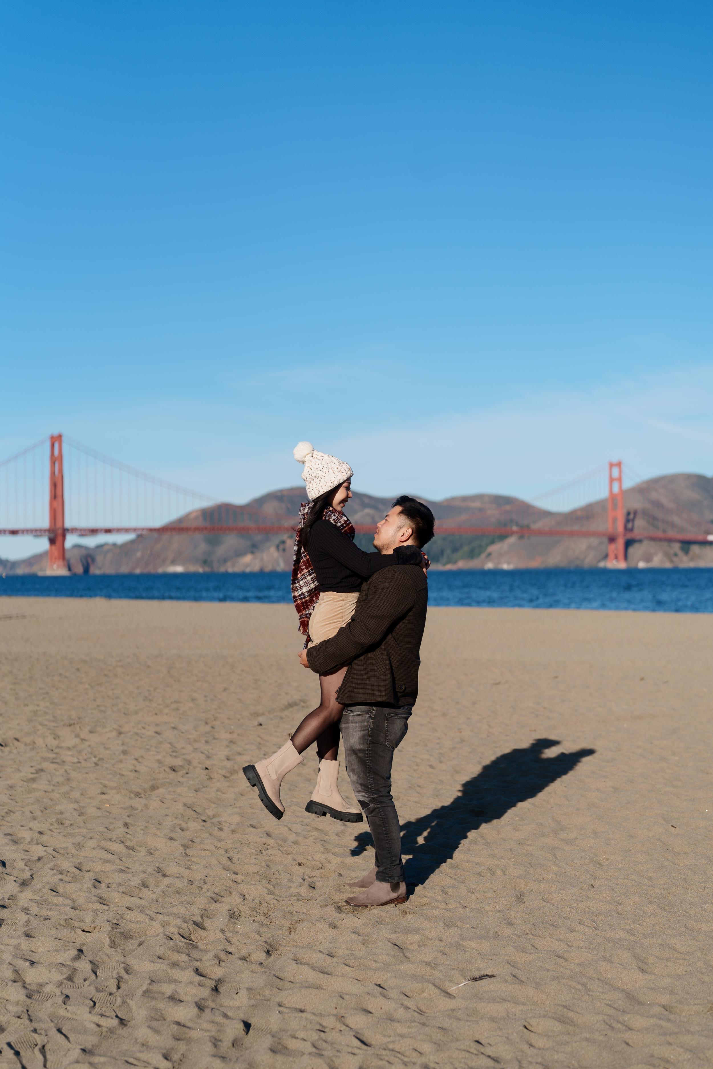 Bay Area couples portrait of a man lifting his partner in a cream beanie and plaid scarf on the sandy beach at Crissy Field