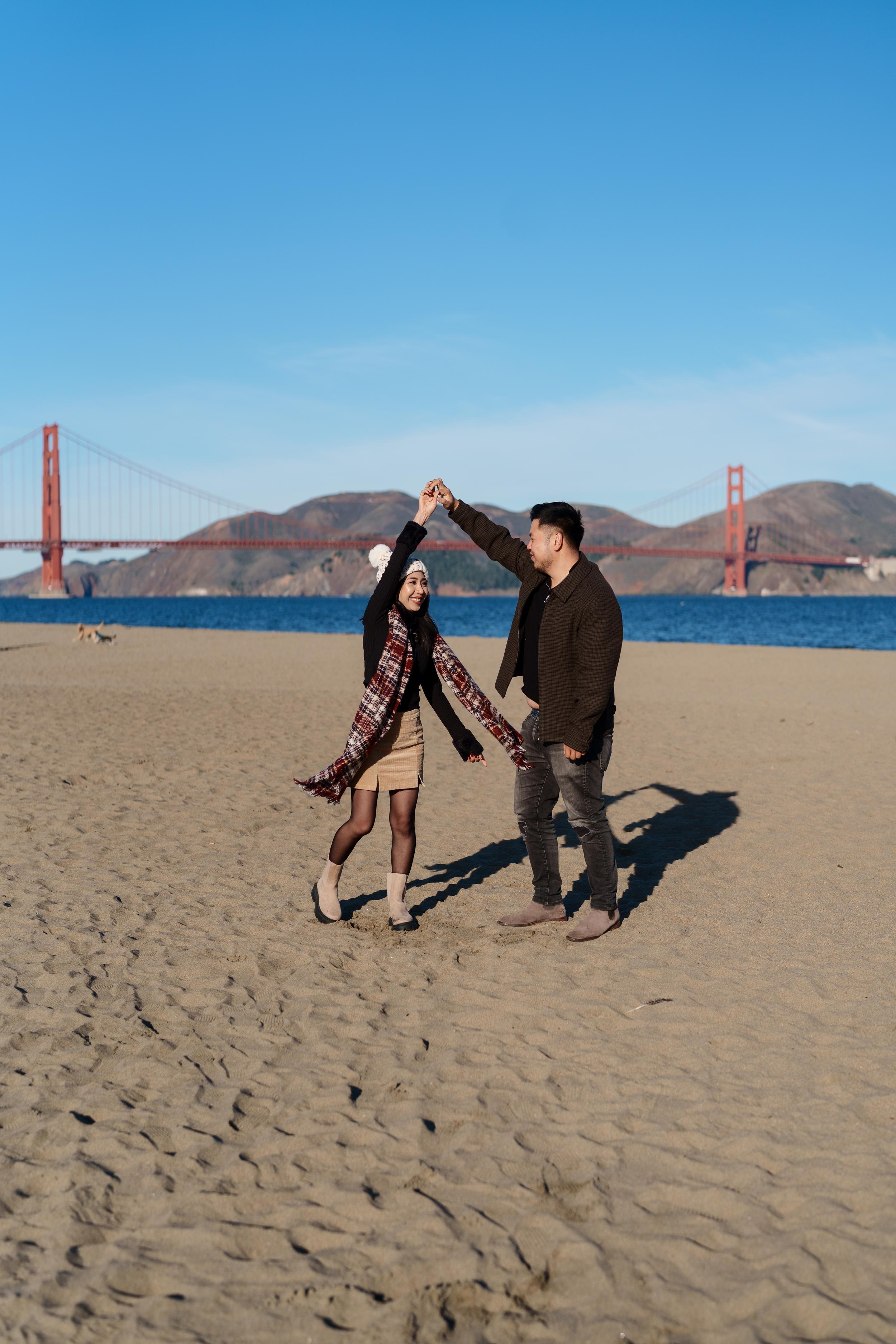 Bay Area couples portrait of a man twirling his partner on the sandy beach at Crissy Field, Golden Gate Bridge behind them in