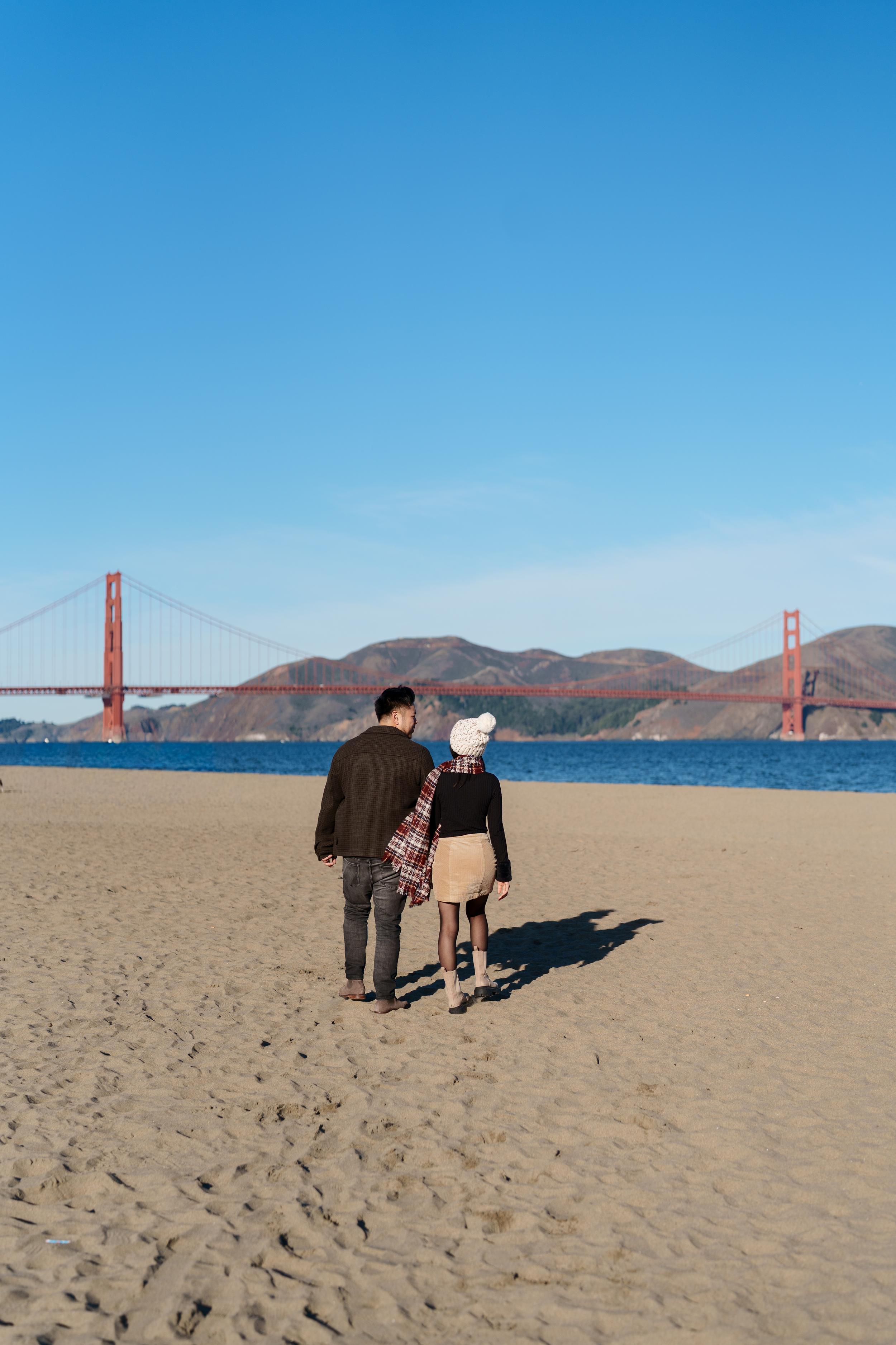 Bay Area couples portrait of a pair walking on Crissy Field beach in cozy fall layers with the Golden Gate Bridge behind