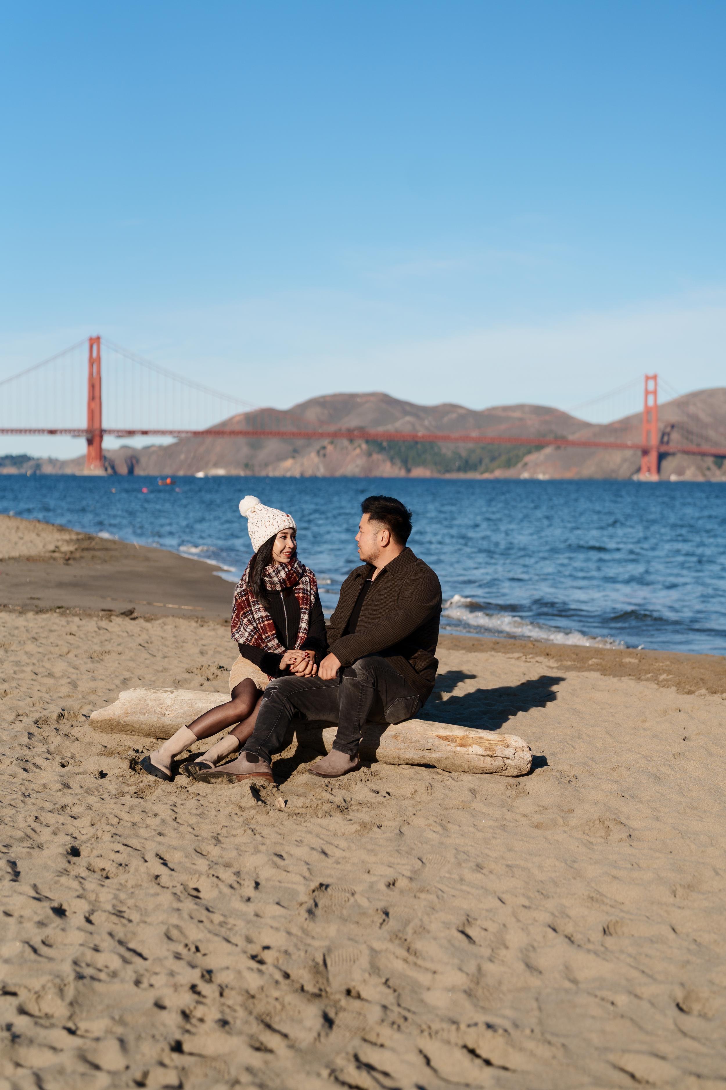 Bay Area couples portrait of two seated on driftwood at Crissy Field beach, Golden Gate Bridge behind them in winter