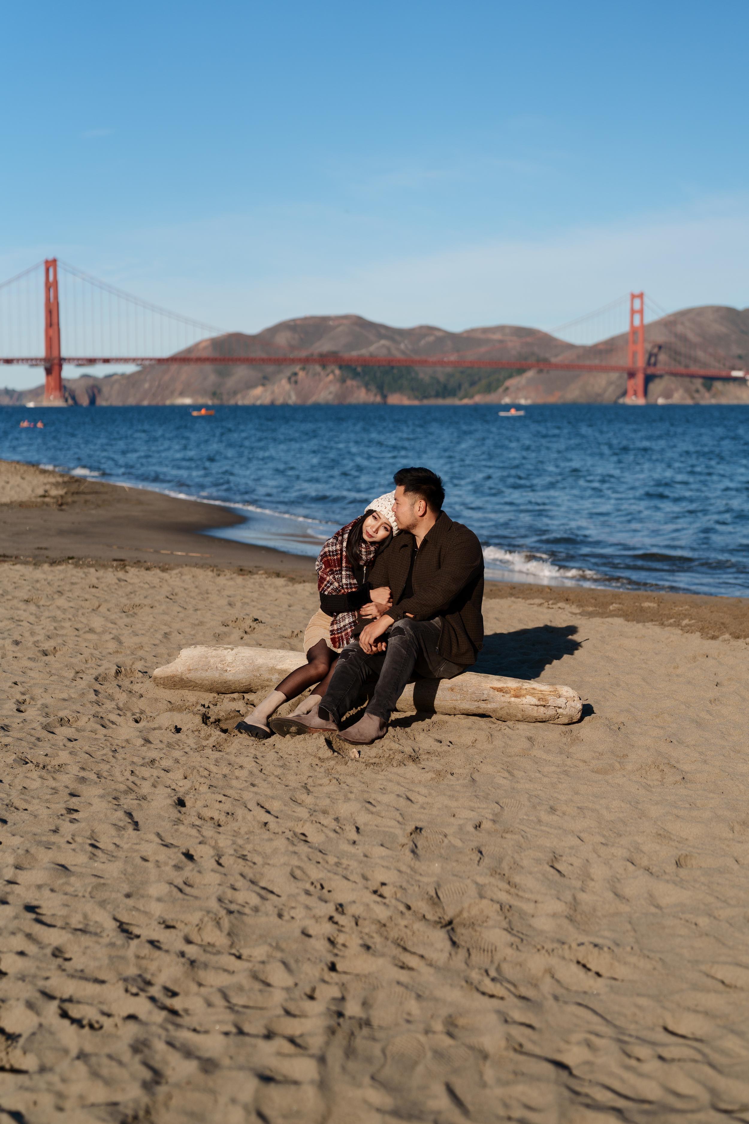 Bay Area couples portrait of a man and woman cuddling on a driftwood log at Crissy Field beach with the Golden Gate Bridge