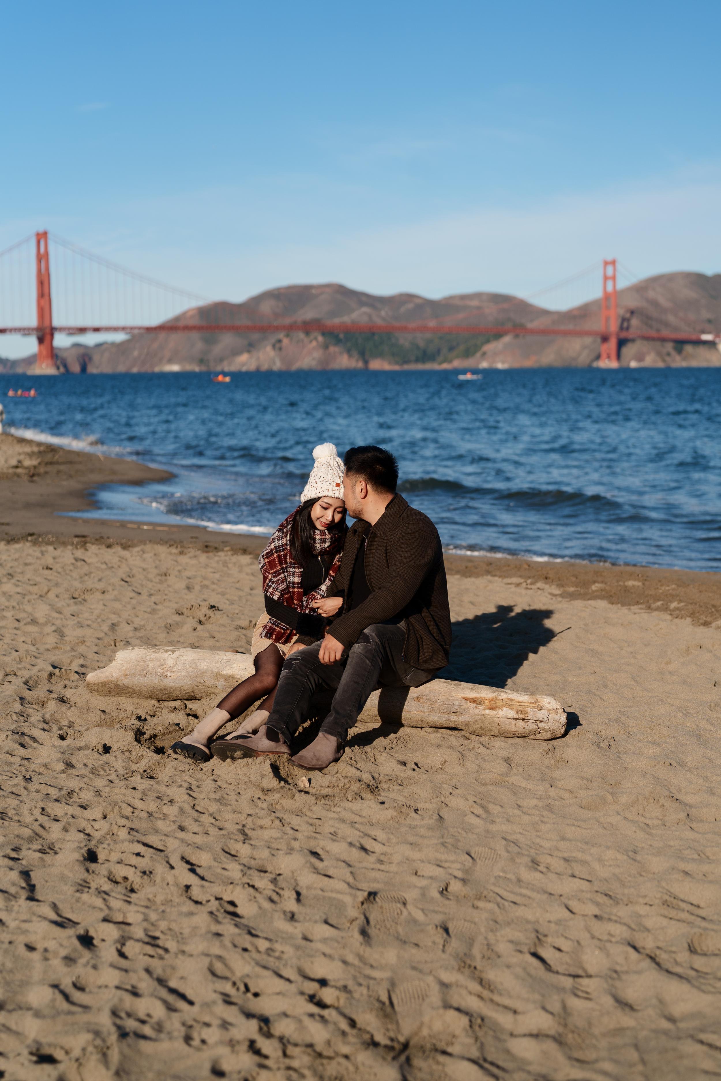 Bay Area couples portrait of a pair sitting on driftwood at Crissy Field beach, Golden Gate Bridge behind them in winter sun.
