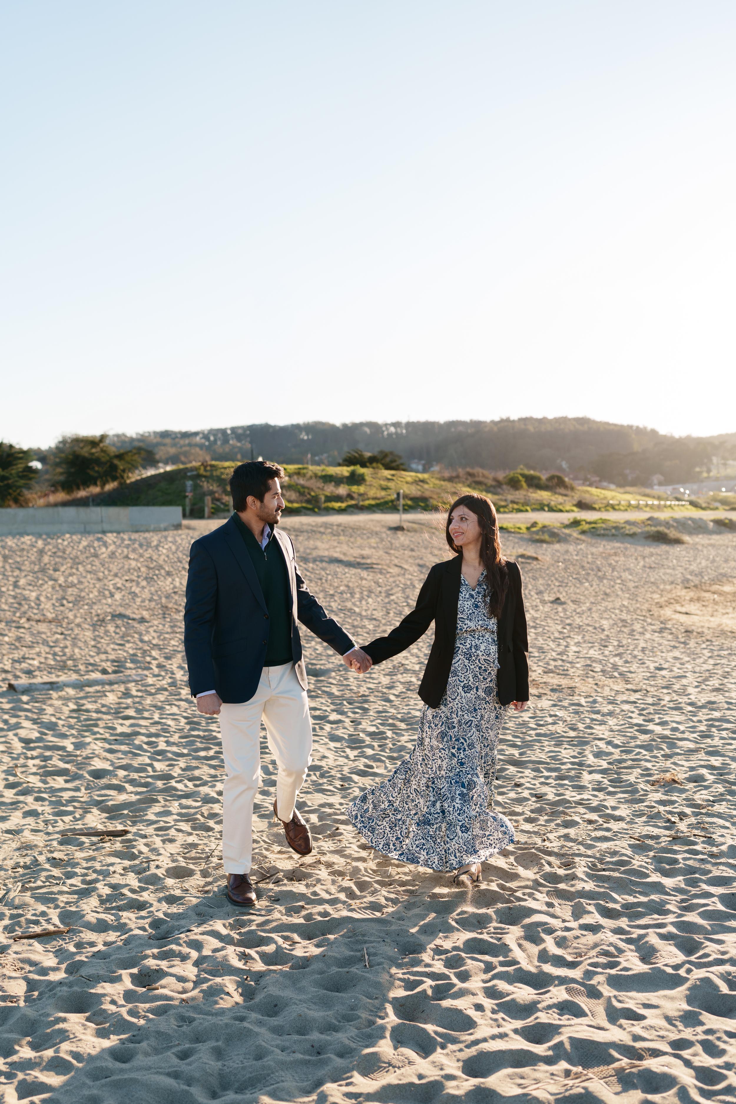 Bay Area couples portrait of a man in navy blazer and woman in blue paisley dress holding hands walking on sandy beach at