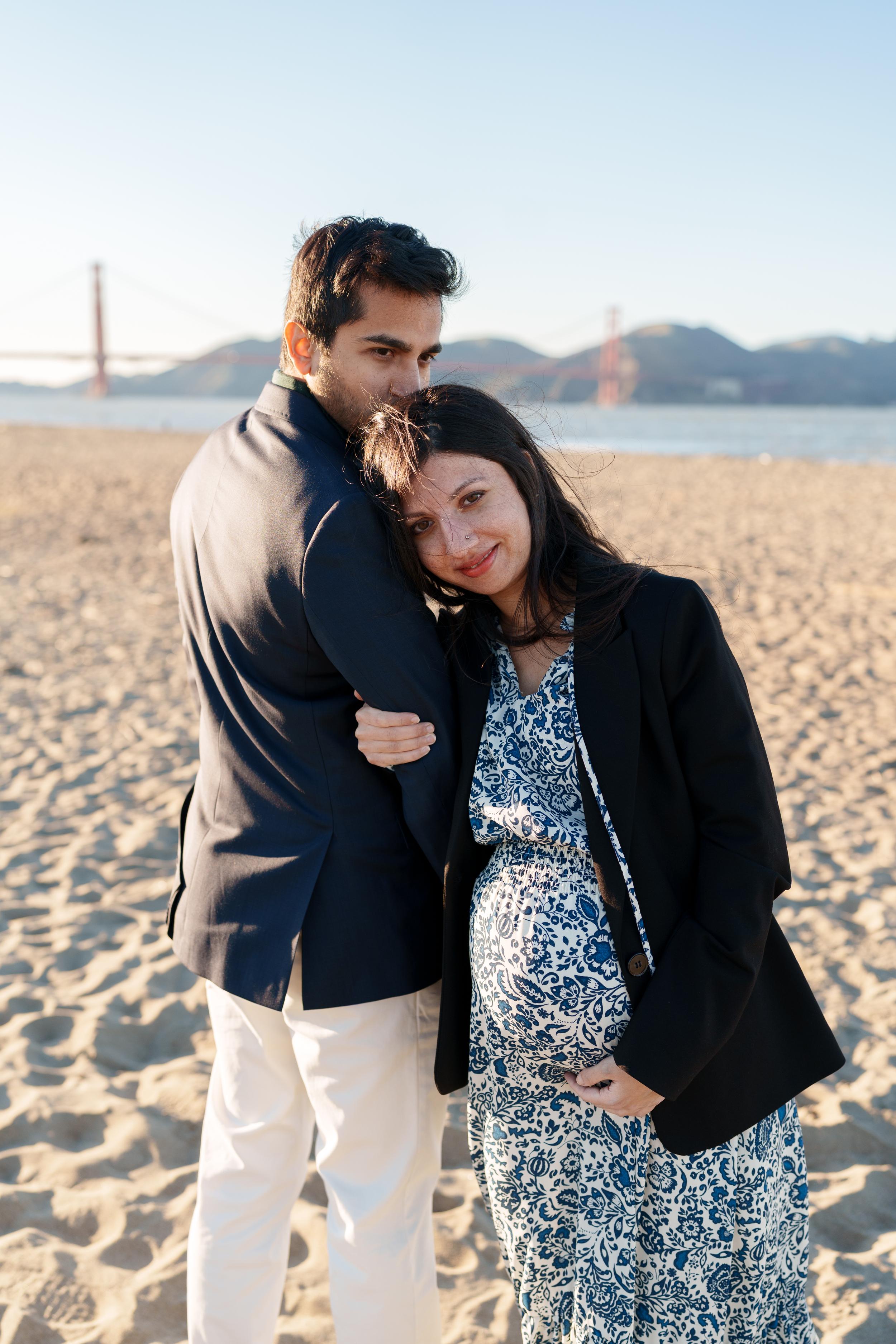 Bay Area couples portrait of expecting parents embracing on Crissy Field sand with Golden Gate Bridge behind them in golden