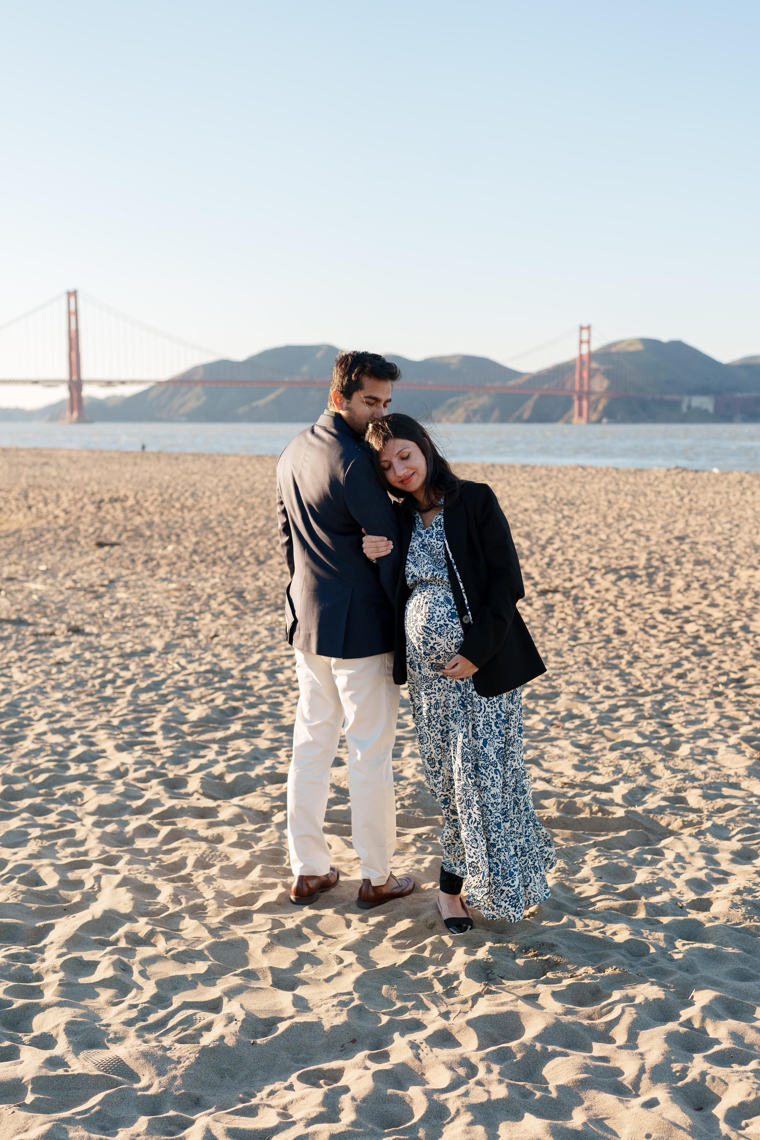 Bay Area couples portrait of expecting parents embracing on sandy Crissy Field beach with the Golden Gate Bridge behind them.