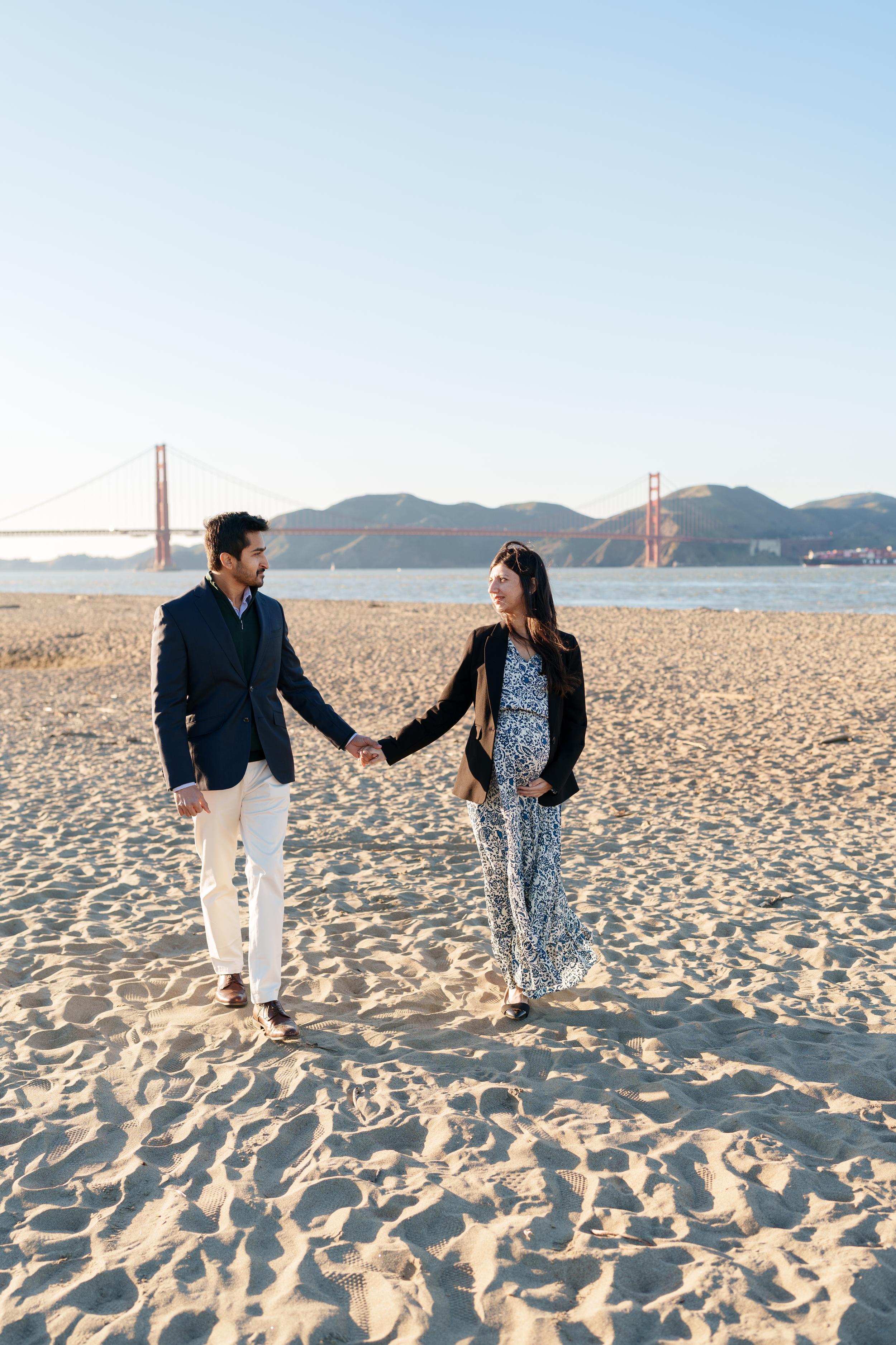 Bay Area couples portrait of an expecting pair holding hands on Crissy Field sand, Golden Gate Bridge behind them.