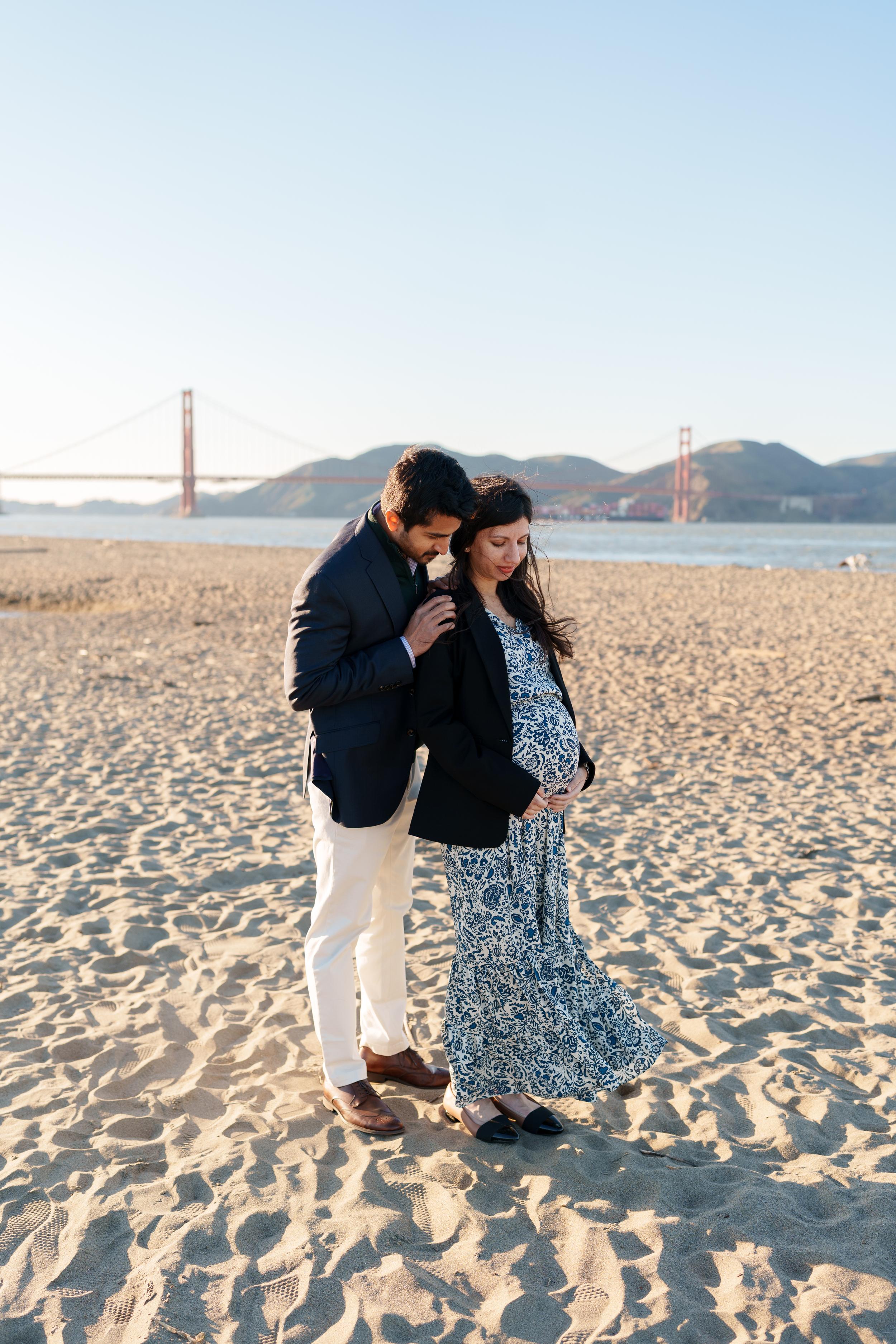 Bay Area couples portrait of expecting partners on sandy Crissy Field beach with Golden Gate Bridge behind them in golden