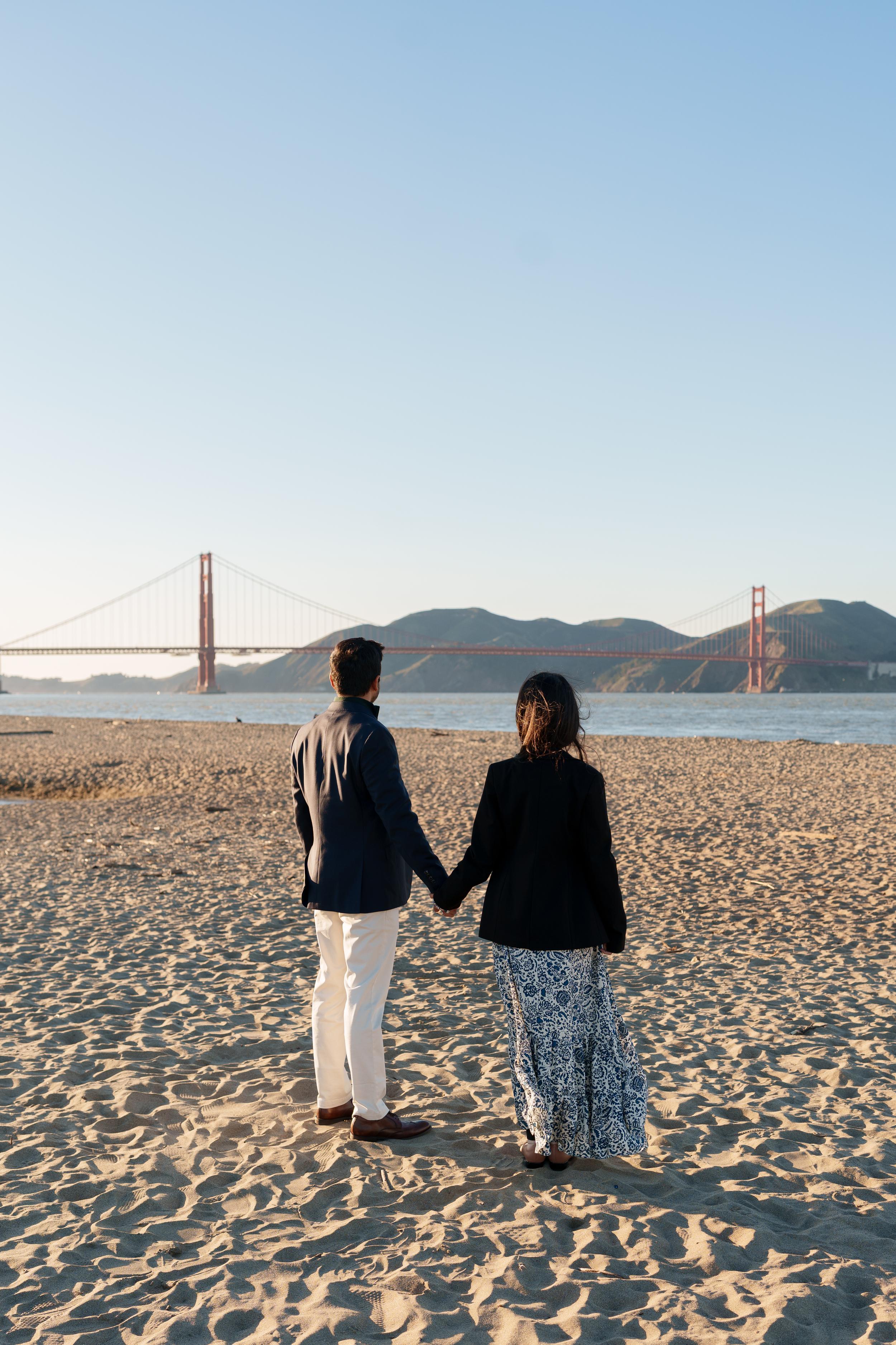 Bay Area couples portrait of a man and woman holding hands on Crissy Field beach, facing the Golden Gate Bridge at sunset.