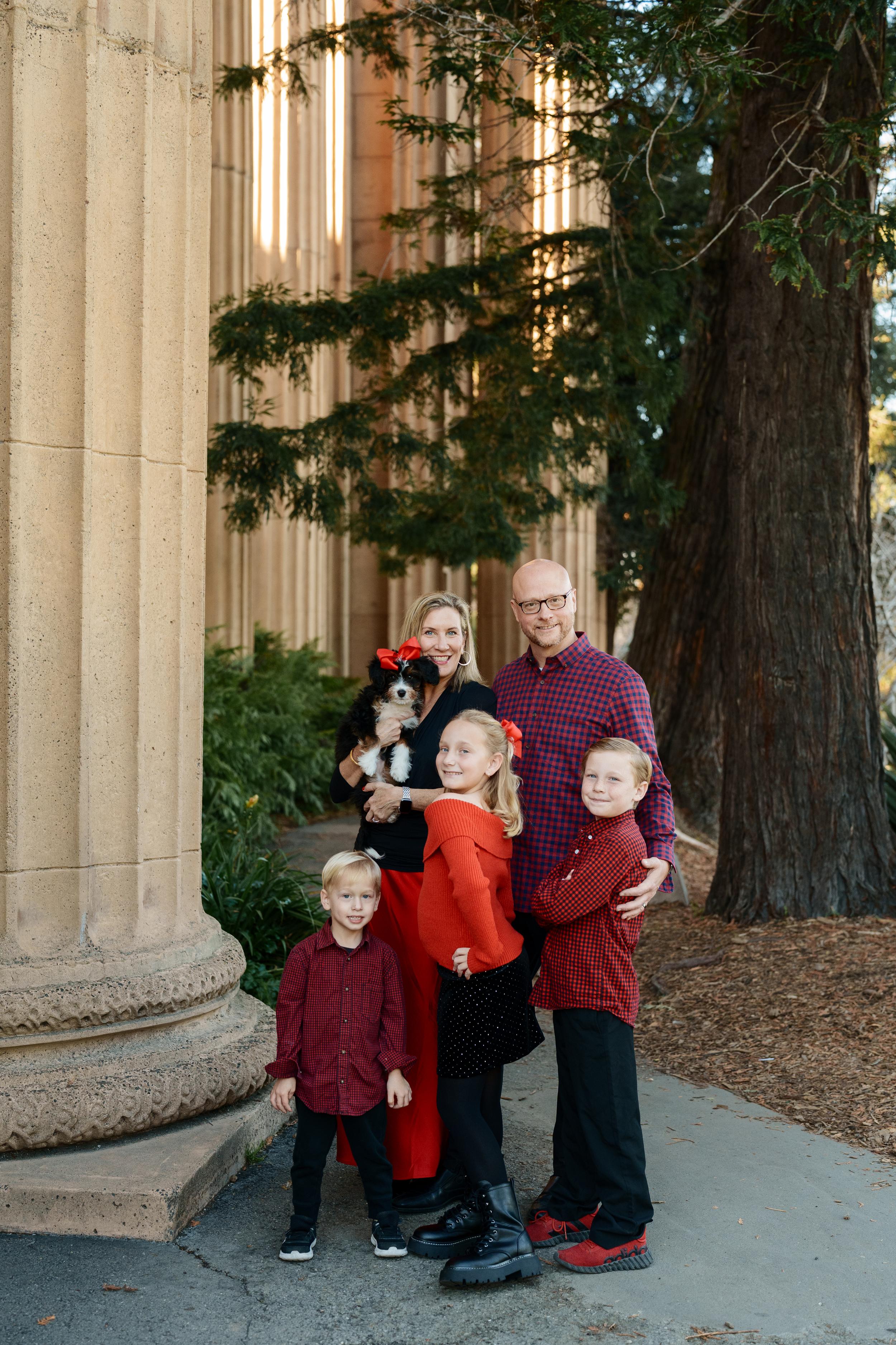 Bay Area family portrait of parents, three kids in red plaid and a puppy with a red bow beside the Palace of Fine Arts