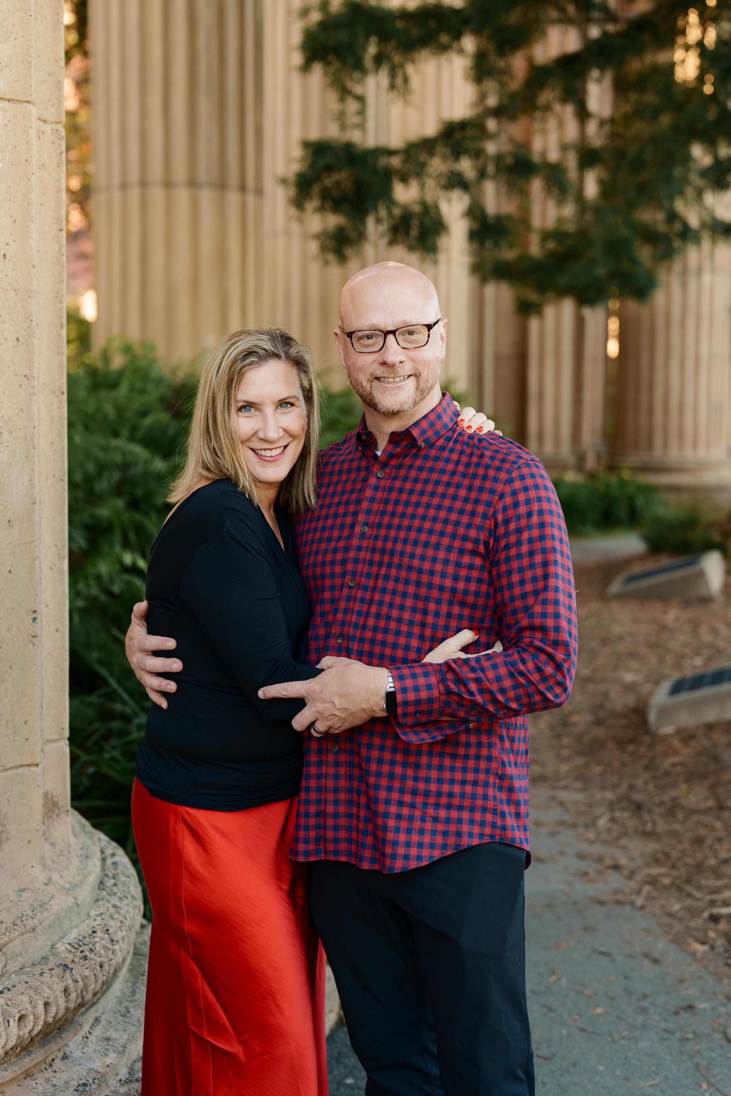 Bay Area family portrait of a smiling couple embracing beside the Palace of Fine Arts columns, woman in black top and red