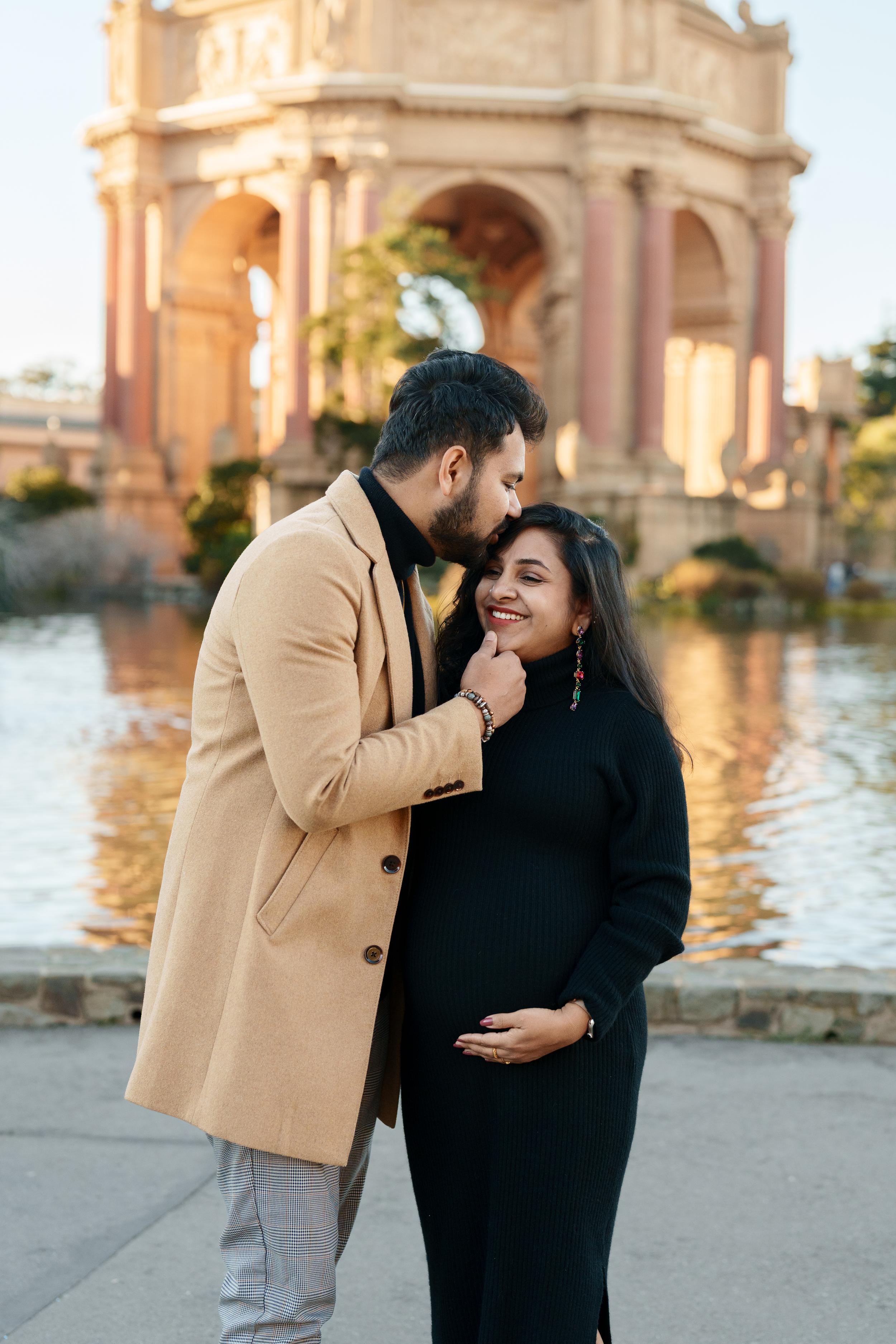 Bay Area couples portrait of expecting parents at Palace of Fine Arts, man in camel coat kissing partner's forehead in black