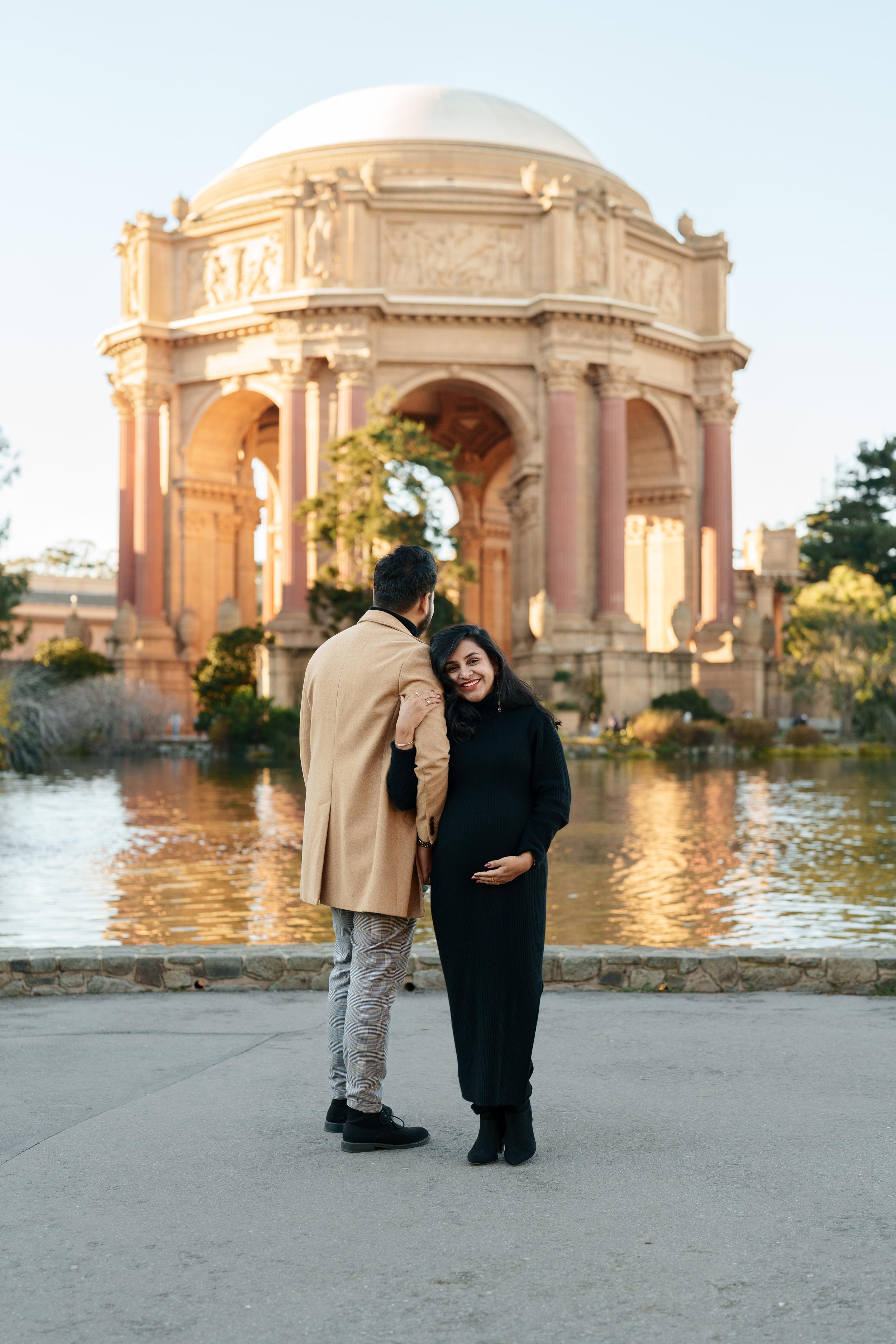 Bay Area couples portrait of an expecting woman in black dress cradling her bump beside partner in tan coat at Palace of Fine