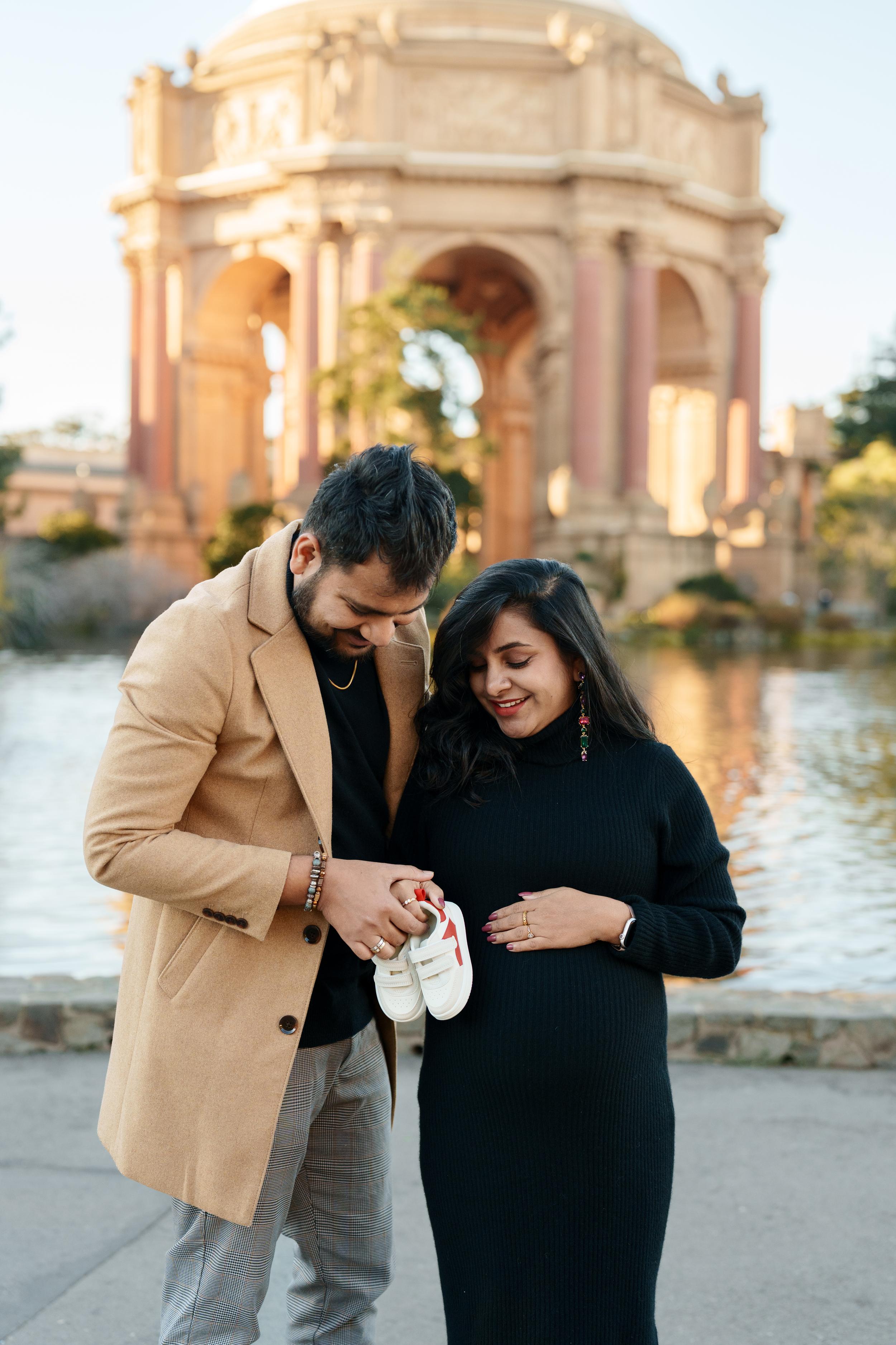 Bay Area couples portrait of expecting parents holding tiny baby sneakers at the Palace of Fine Arts at golden hour.