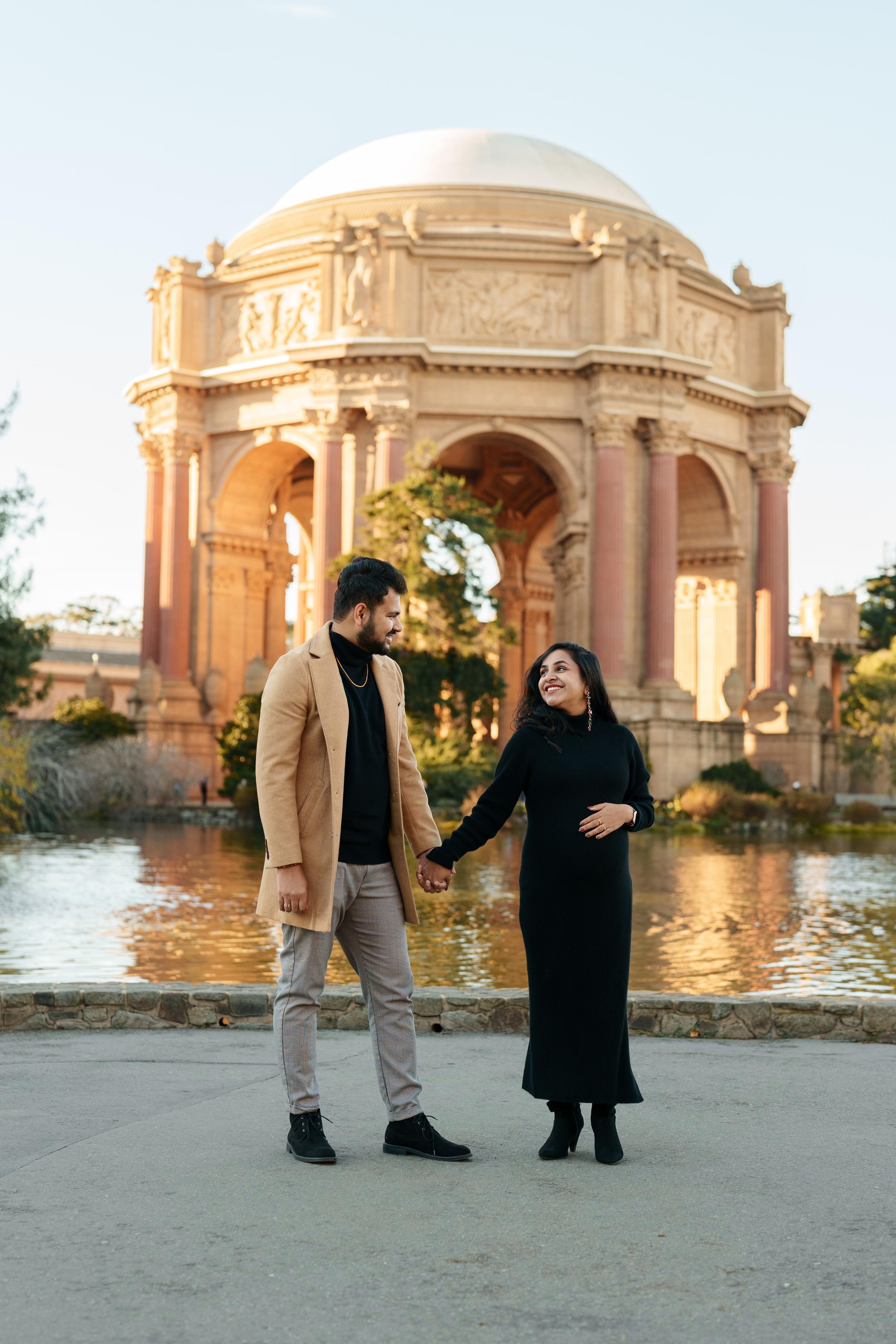 Bay Area couples portrait of a smiling pair holding hands by the lagoon, man in camel coat and woman in black dress at the
