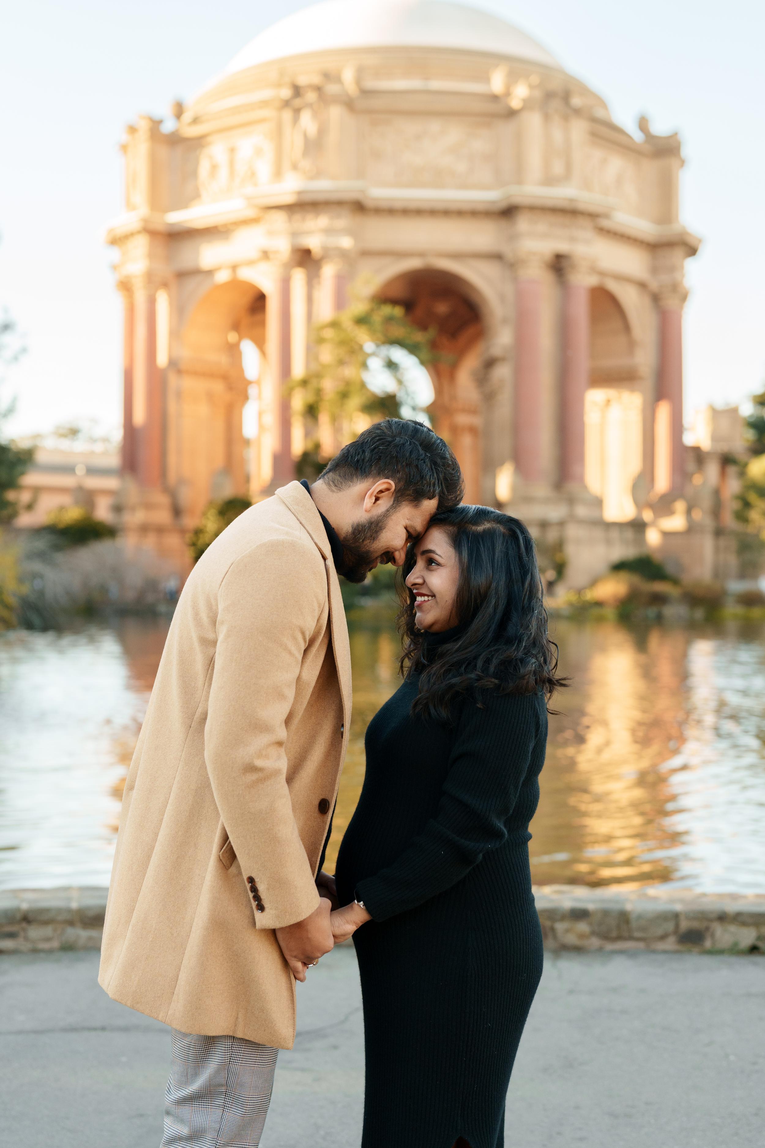 Bay Area couples portrait of a man in camel coat and woman in black dress touching foreheads at Palace of Fine Arts lagoon.