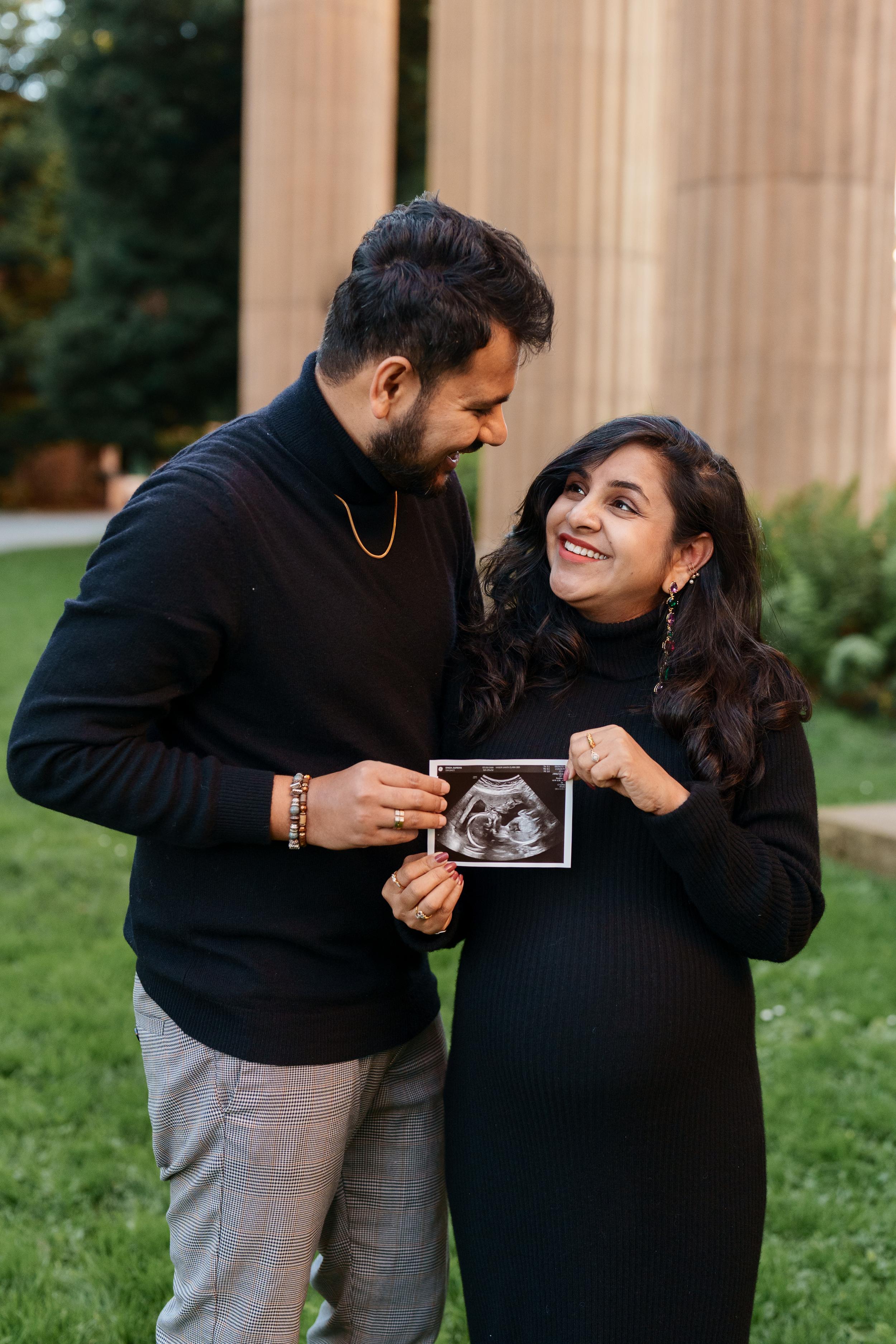 Bay Area couples portrait of expecting parents in black turtlenecks holding an ultrasound photo near tall wooden columns at