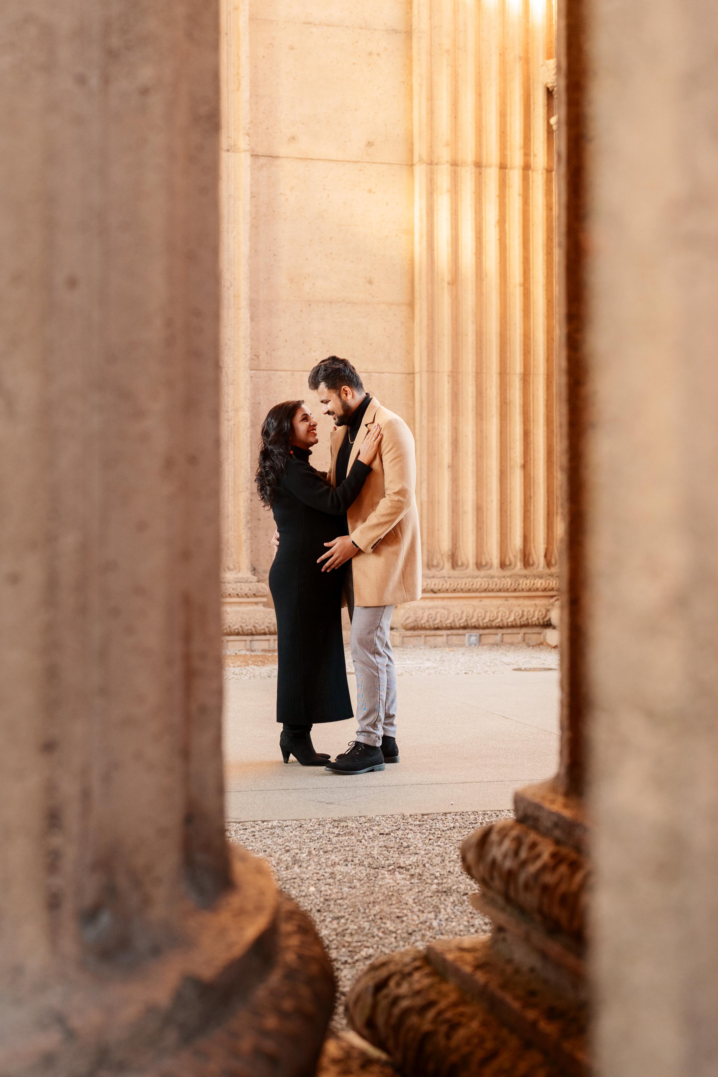 Bay Area couples portrait of a pair embracing between sandstone columns at the Palace of Fine Arts, she in a black dress, he