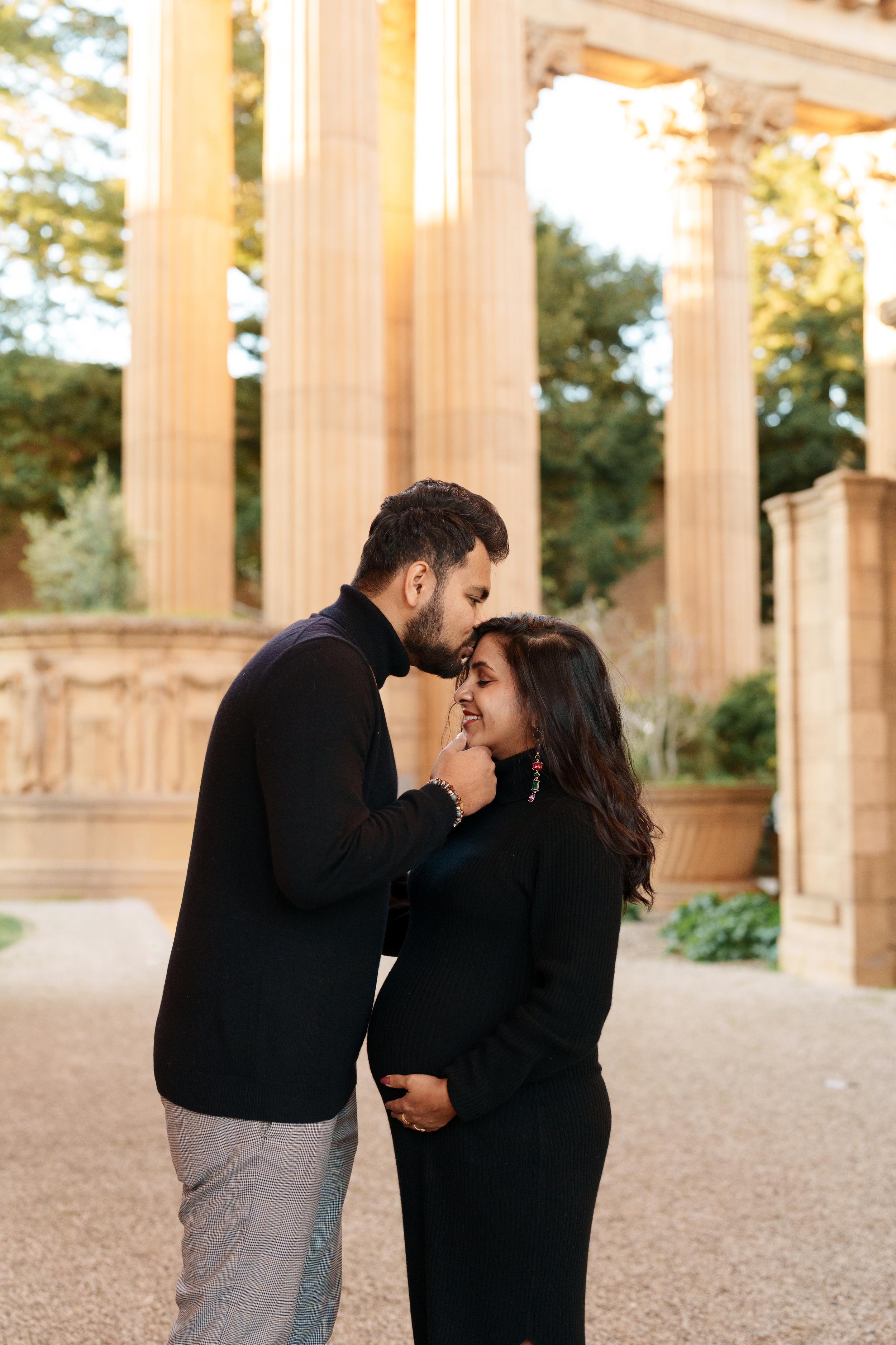 Bay Area couples portrait of expecting parents in black turtlenecks embracing as he kisses her forehead beneath sunlit