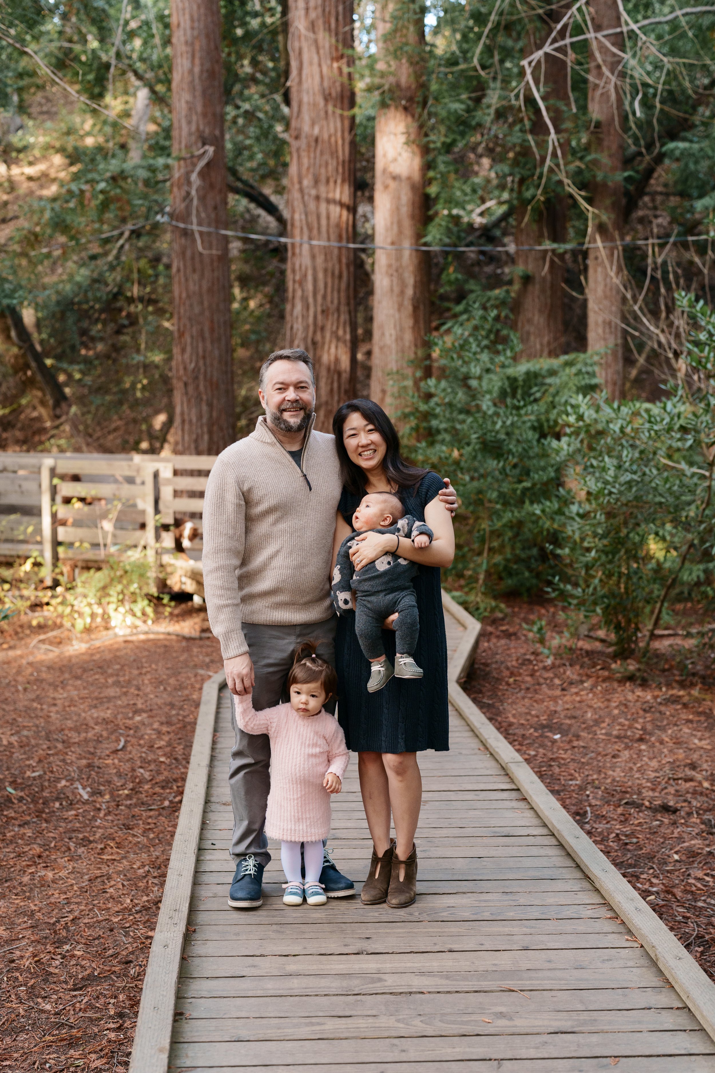 Bay Area family portrait of parents with toddler in pink dress and baby in koala outfit on a wooden boardwalk among redwoods.