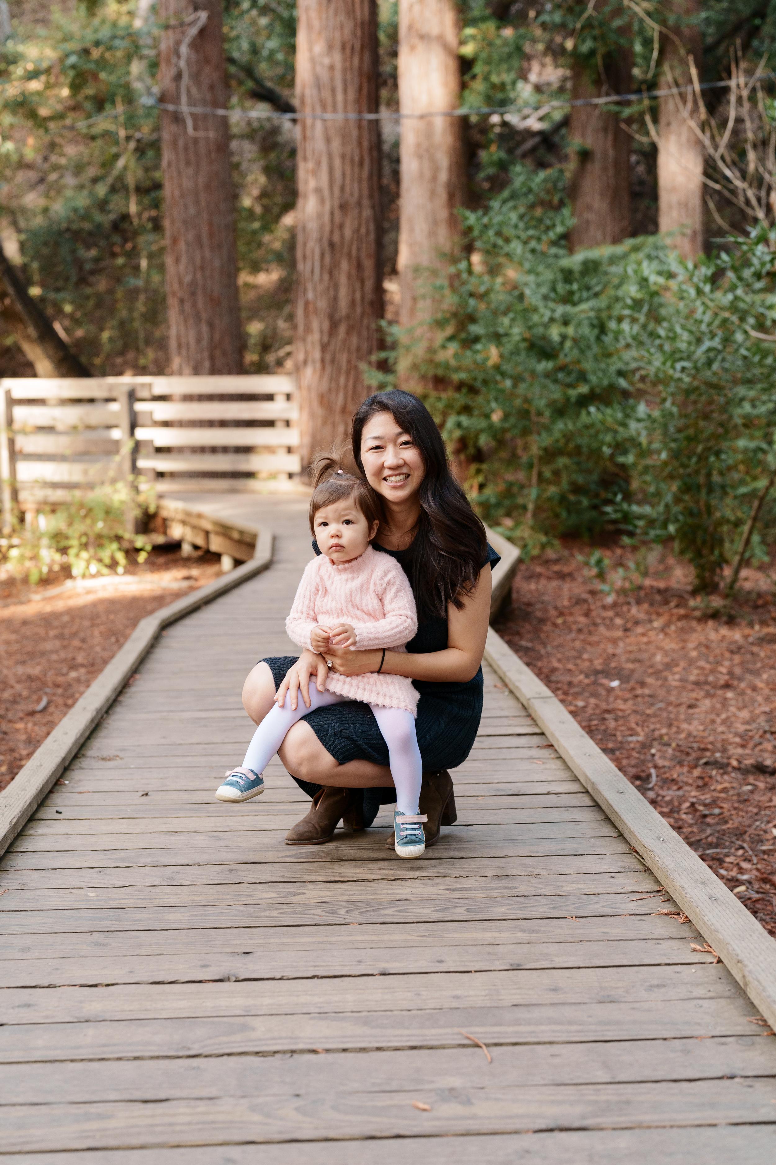 Bay Area family portrait of a smiling mom in navy dress crouching with her toddler daughter in pink sweater on a redwood
