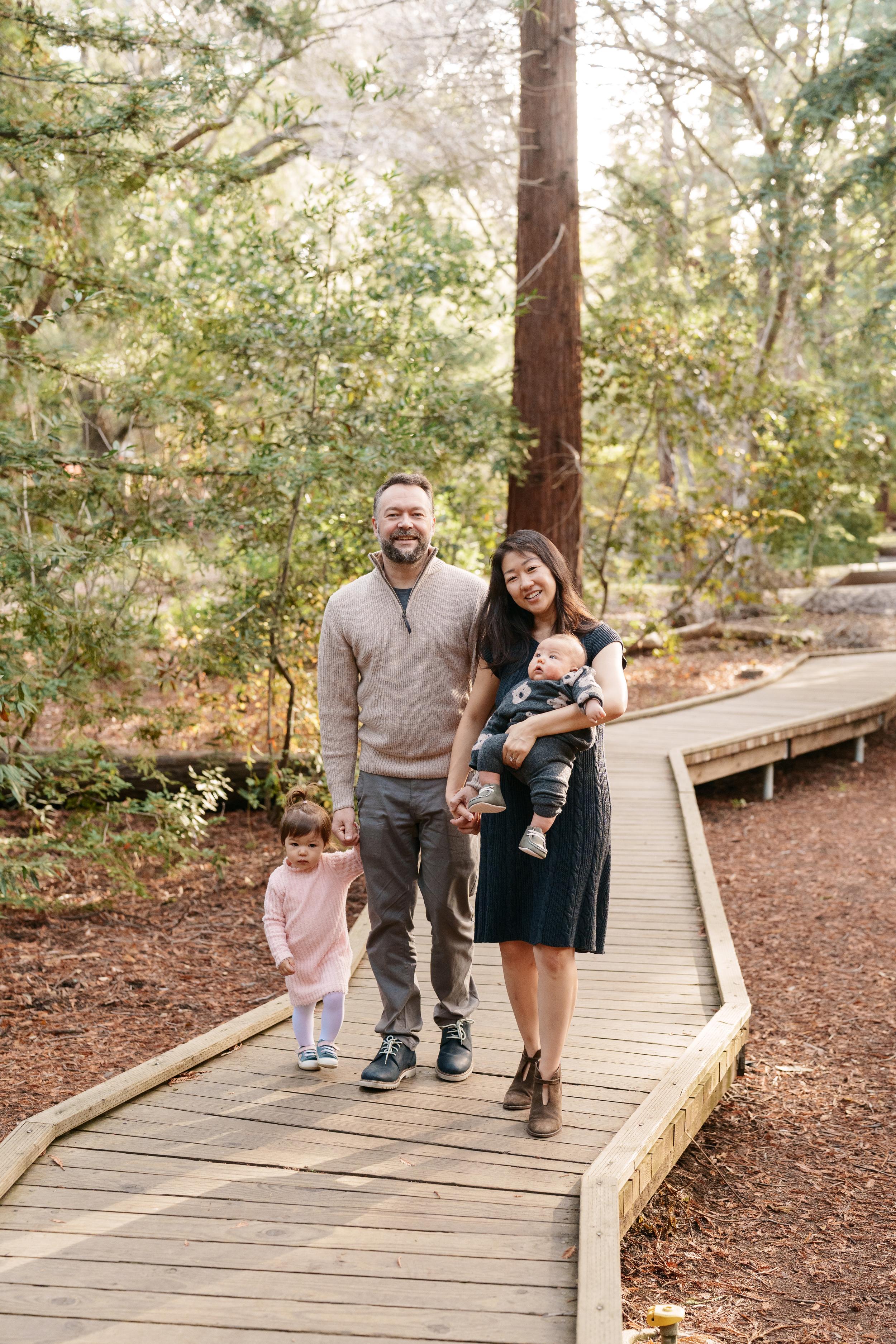 Bay Area family portrait of parents with toddler in pink dress and baby boy on a wooden boardwalk among redwood trees.